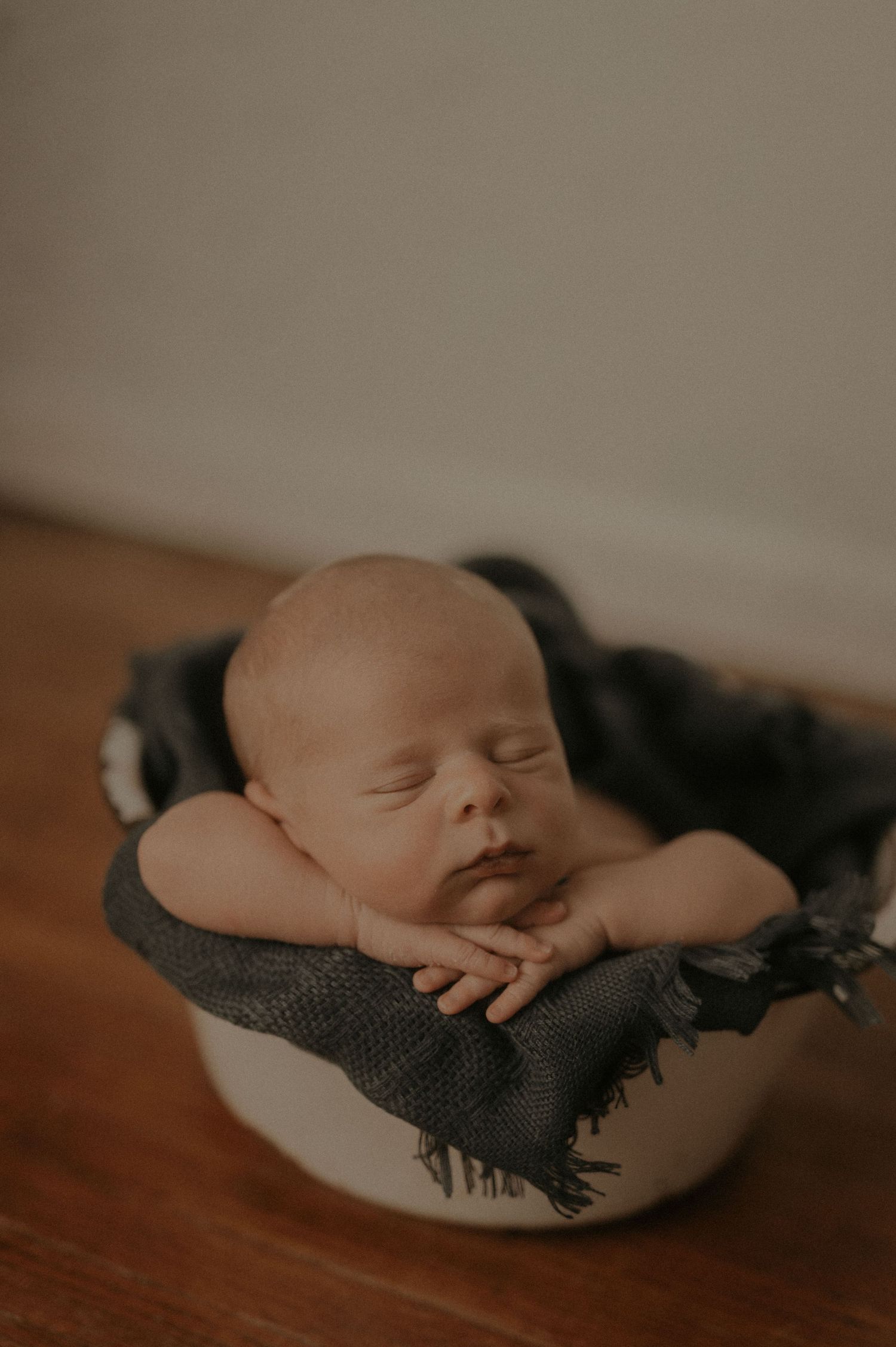 Sleeping newborn nestled in gray fabric inside white bowl on wooden surface.