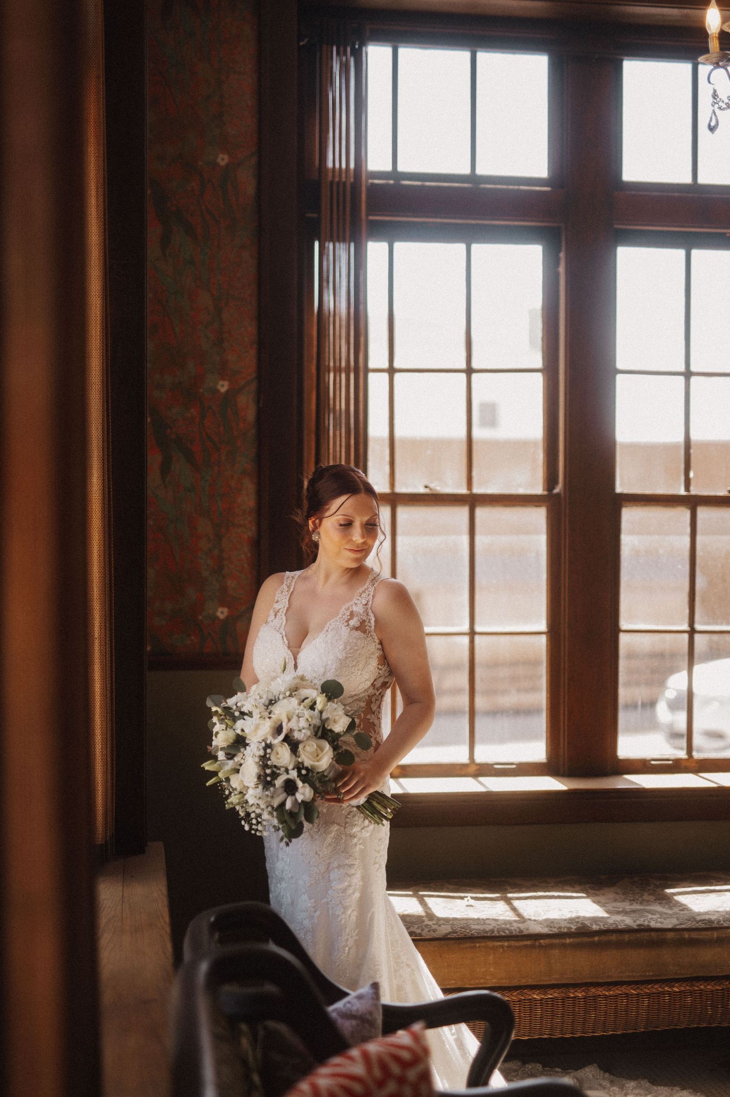 A bride in a white lace gown stands by a window in a vintage room with warm natural lighting.