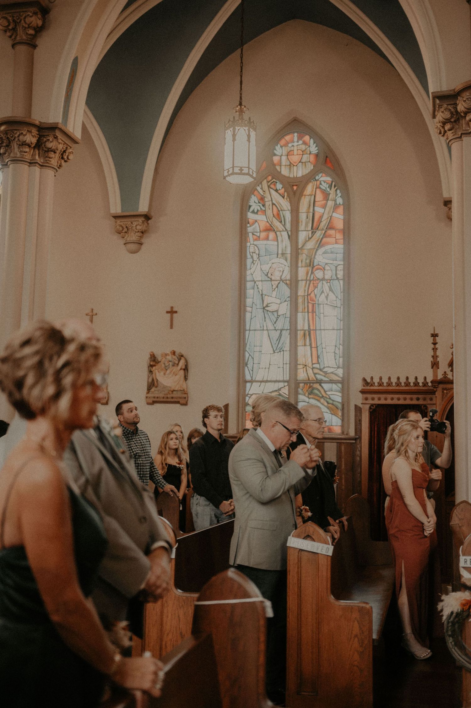 Church interior with stained glass windows and attendees standing in wooden pews during a ceremony.