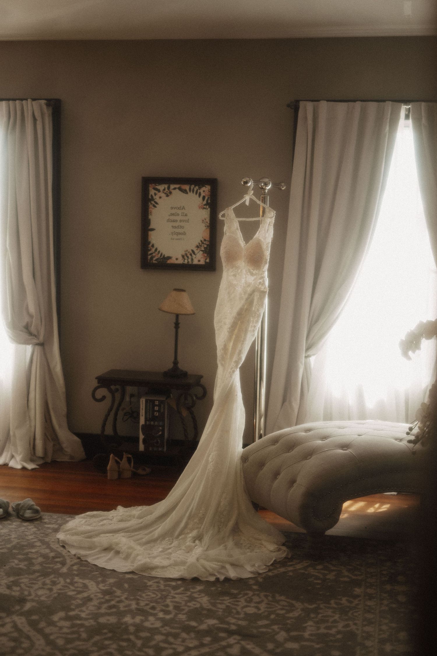 A wedding dress hangs elegantly against white curtains in a softly lit room.