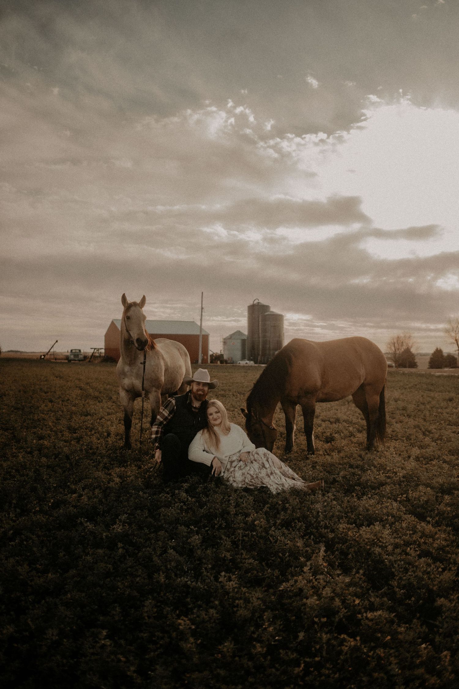 Couple sitting in a field with two horses while dramatic clouds hover over a distant cityscape.