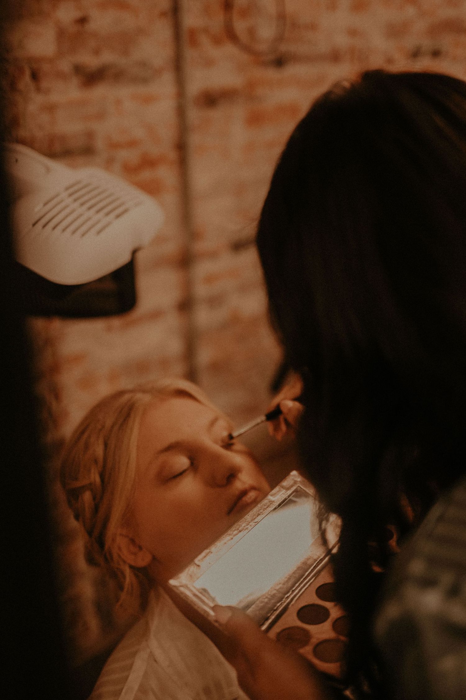A makeup artist applies cosmetics in dramatic lighting against exposed brick walls.