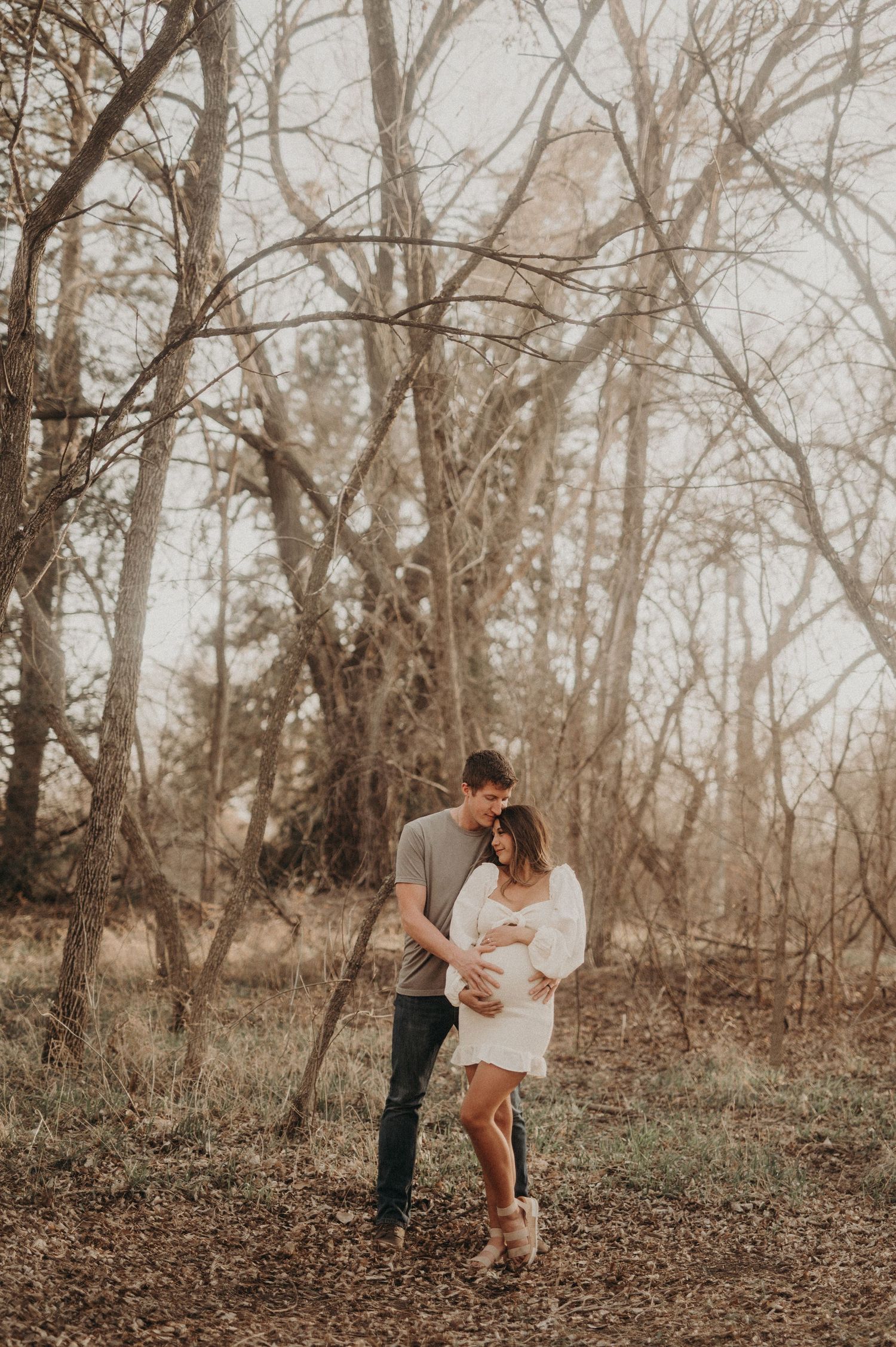 Romantic couple embraces during maternity photoshoot in a wooded forest setting.