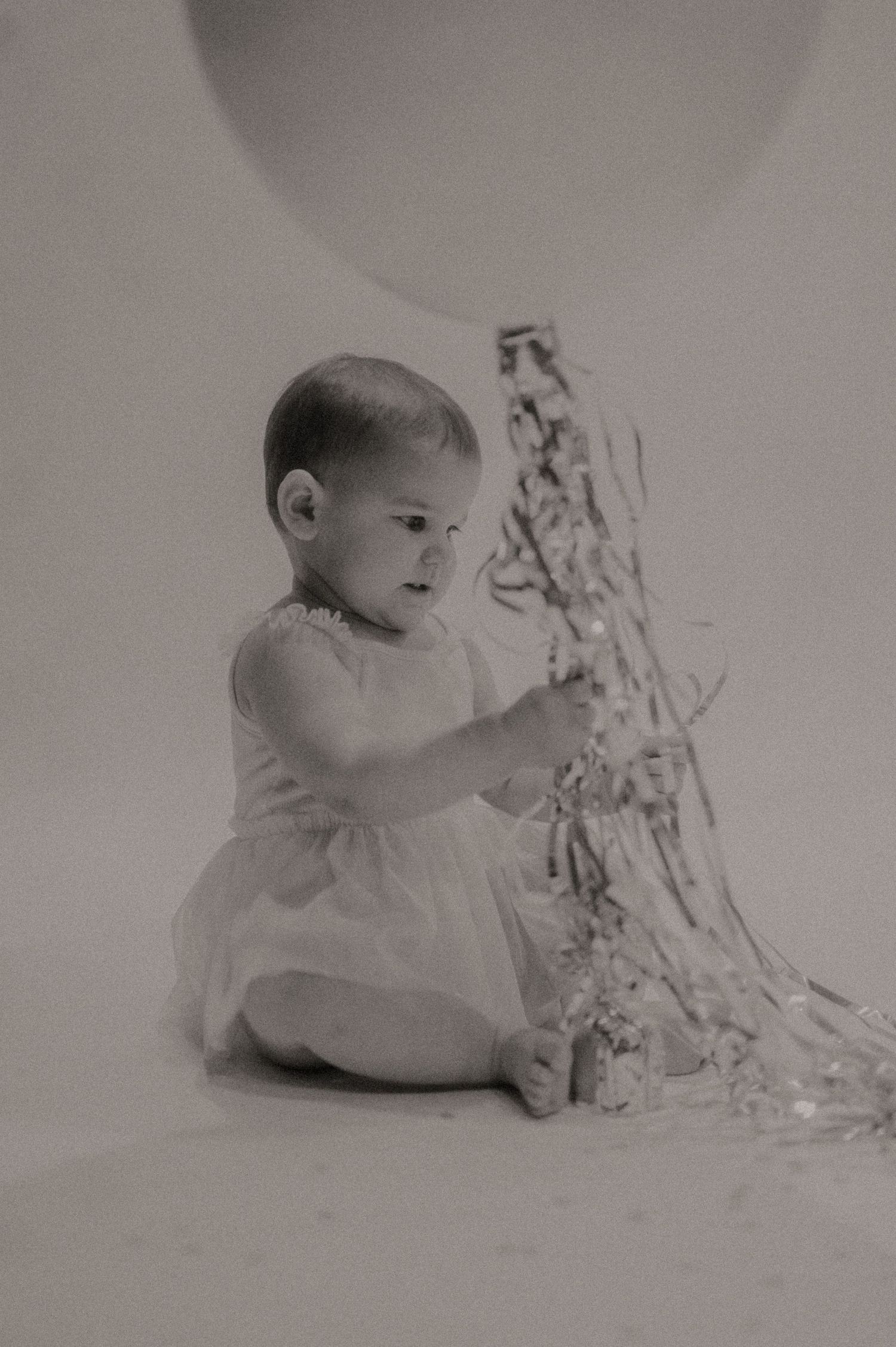A baby in white clothing explores a white floral branch against a light background in a vintage black and white photo.
