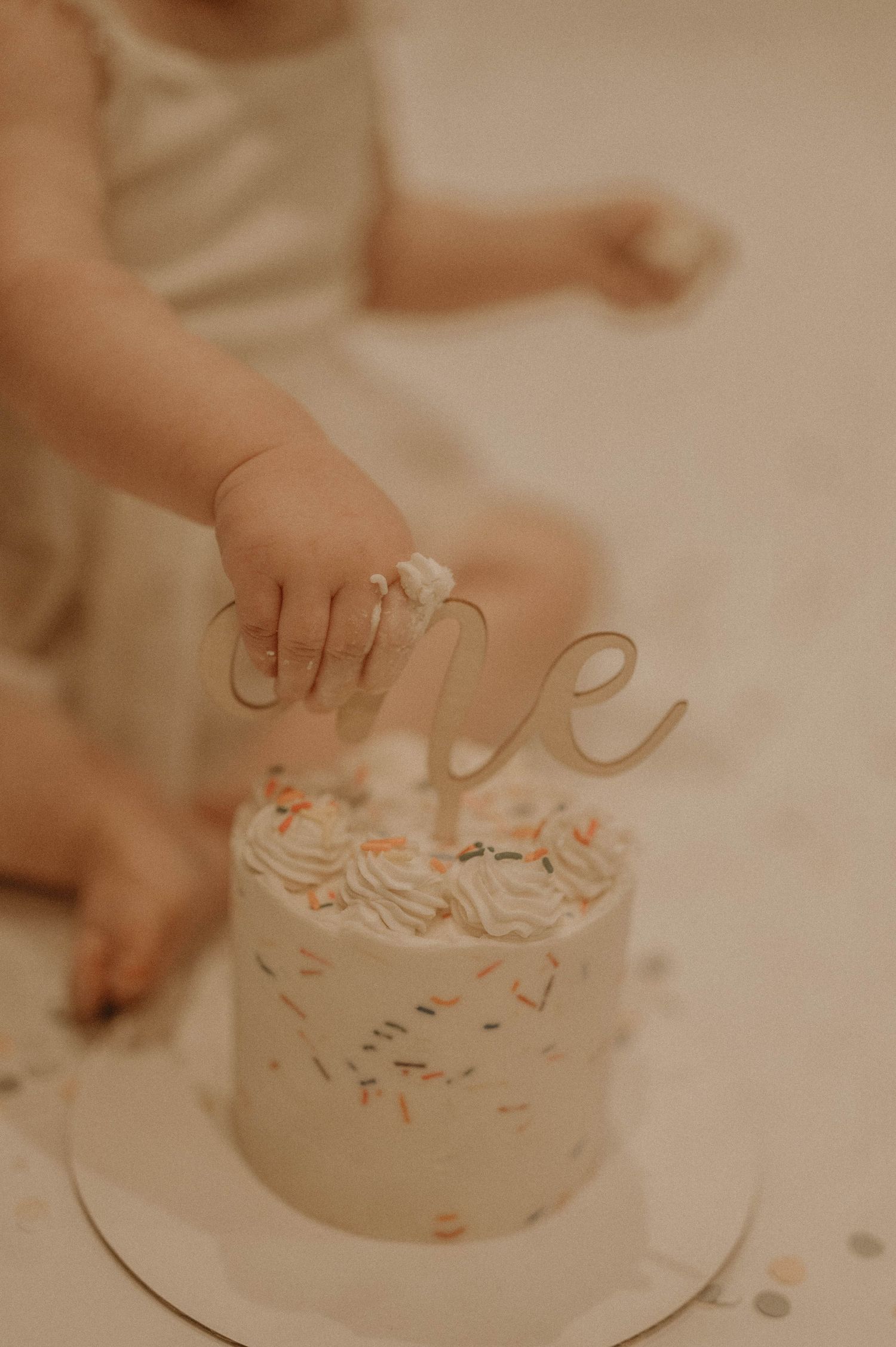 Small hands reaching to touch a white frosted birthday cake with delicate floral decorations on a light beige background.