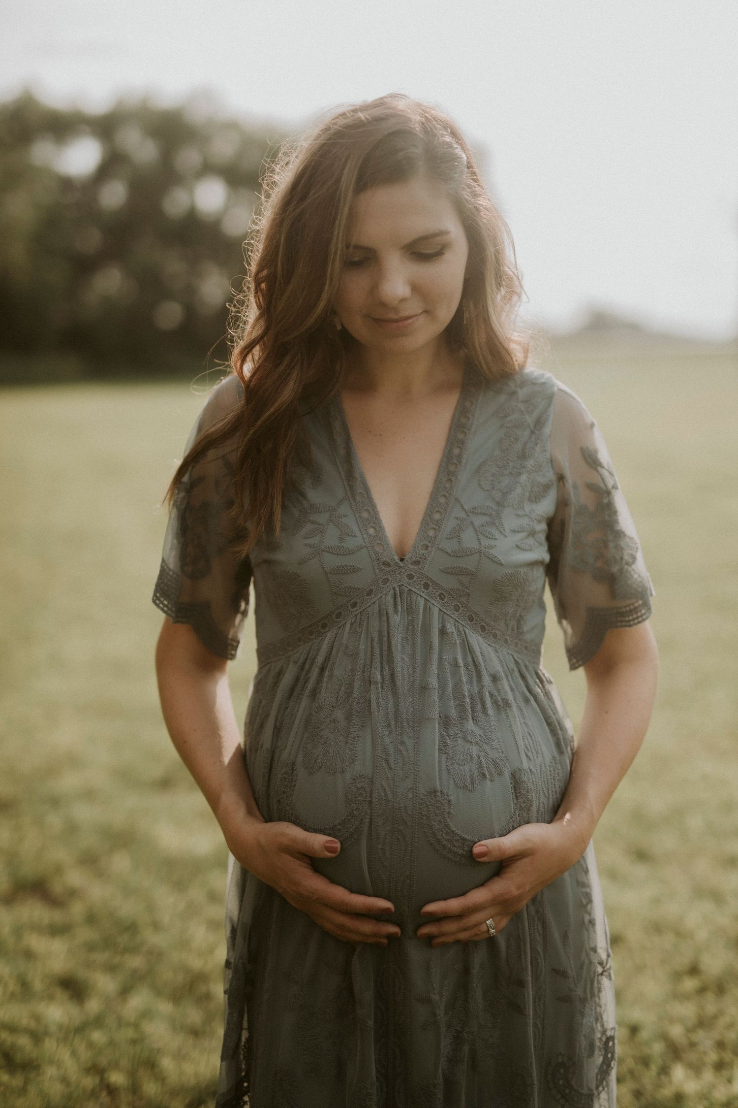 A pregnant woman in a green dress stands in a sunlit field during a maternity photoshoot.