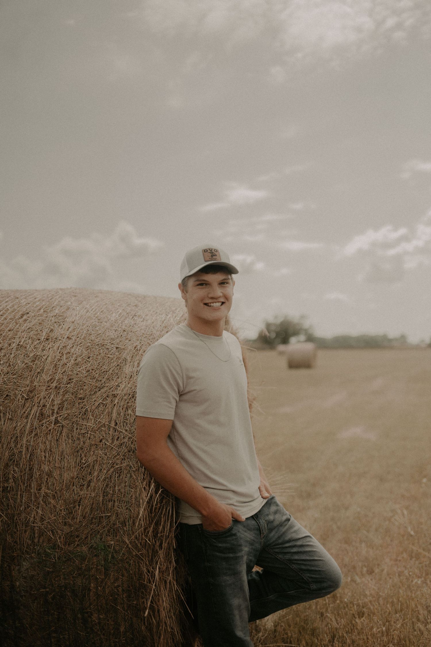 A person in a baseball cap and t-shirt leans against a hay bale in a rural field during golden hour.