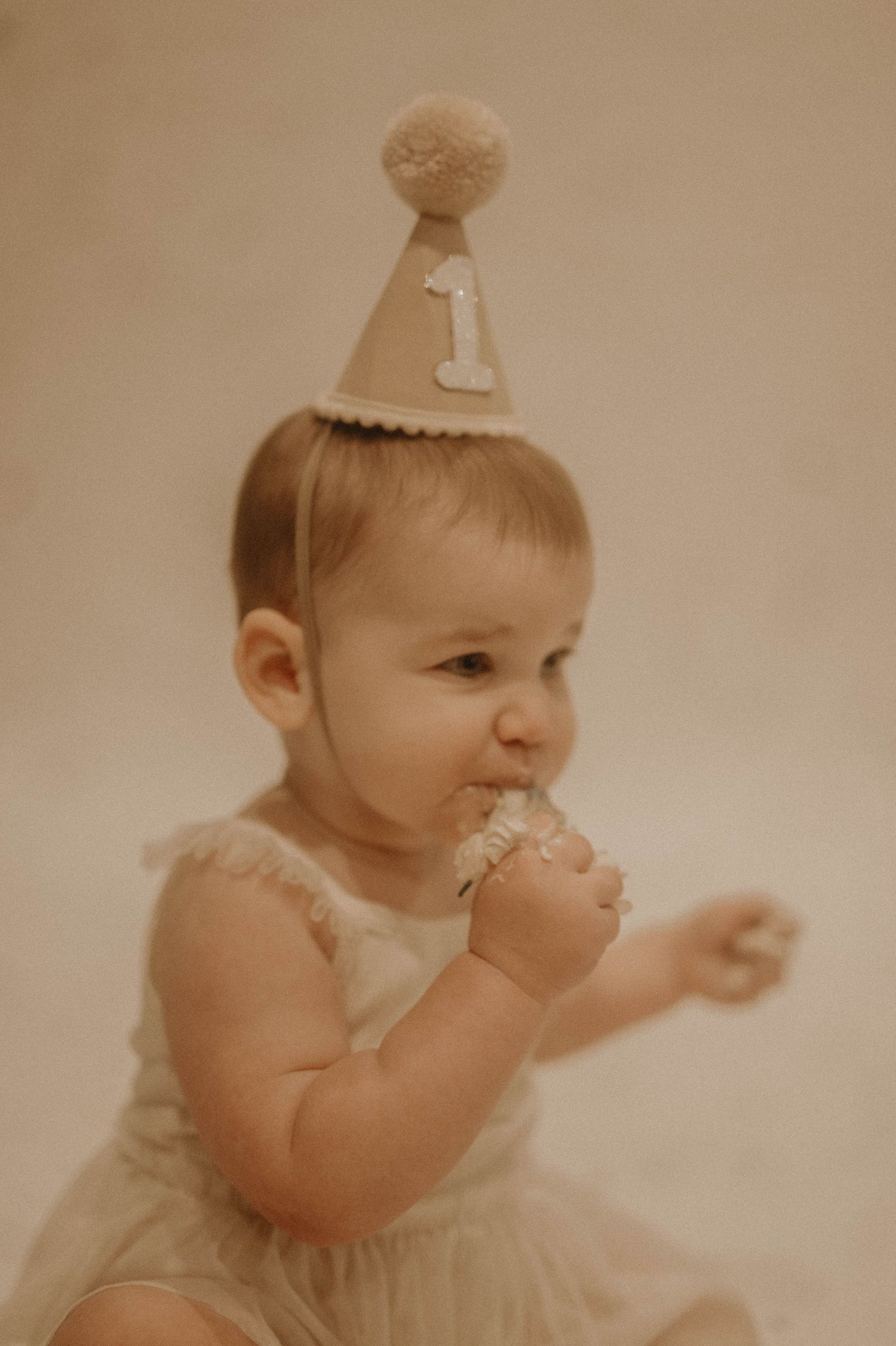 A baby in a white outfit with a birthday hat celebrates their first birthday by eating cake.