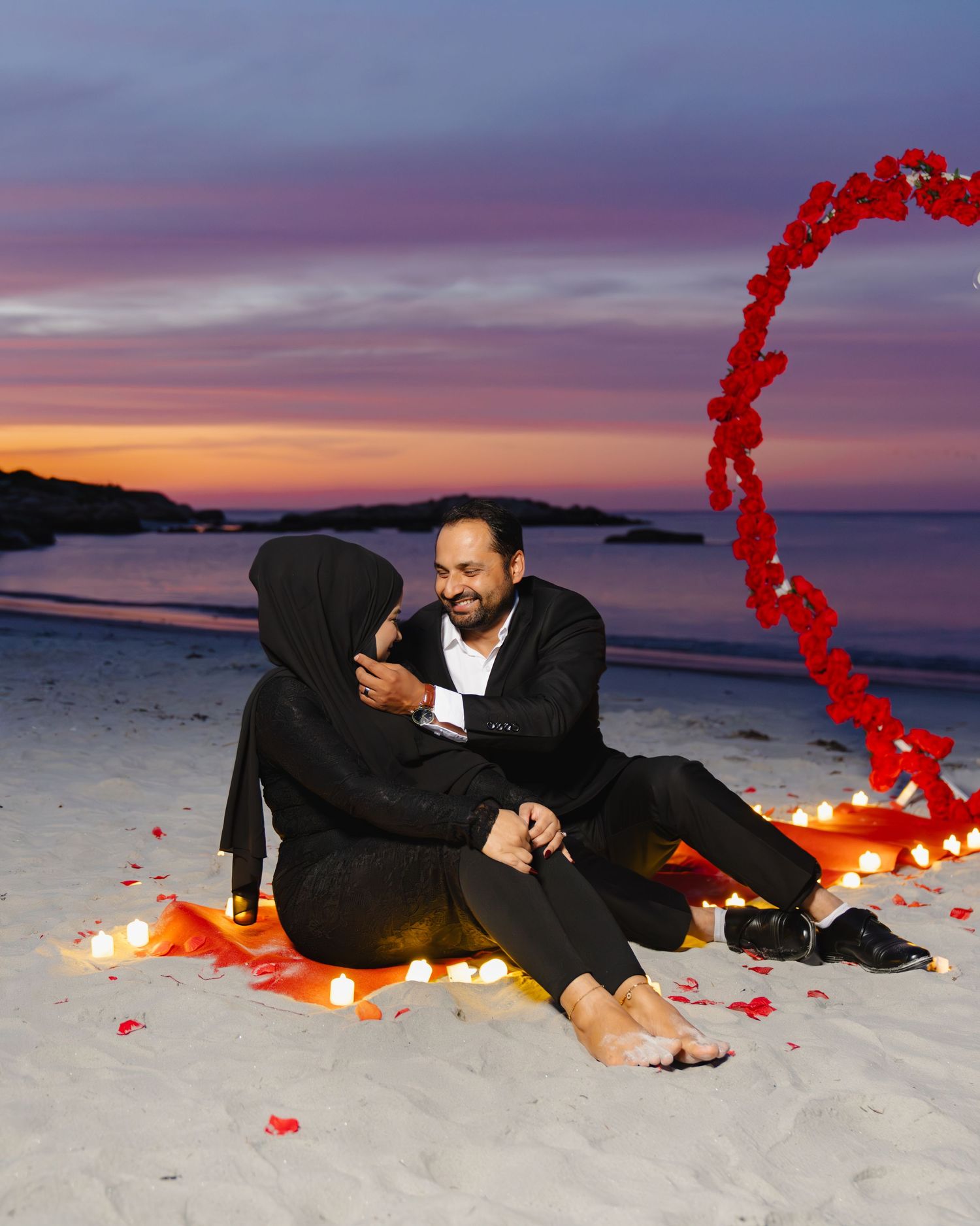 A couple lies on a beach at sunset with a trail of glowing red rose petals creating a heart shape around them. Boston MA photographer.