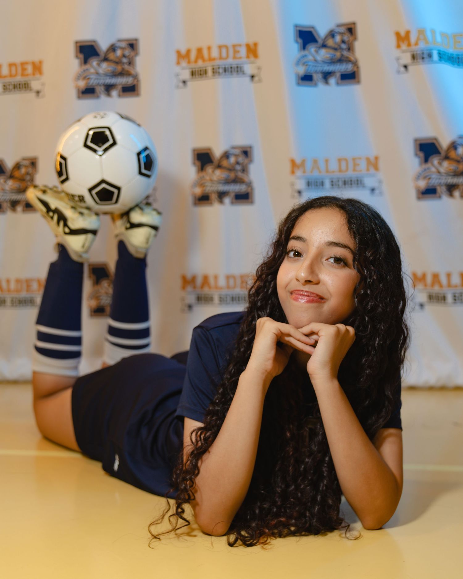 A person with curly dark hair lies on their stomach in front of a branded backdrop while balancing a soccer ball. Boston MA photographer.