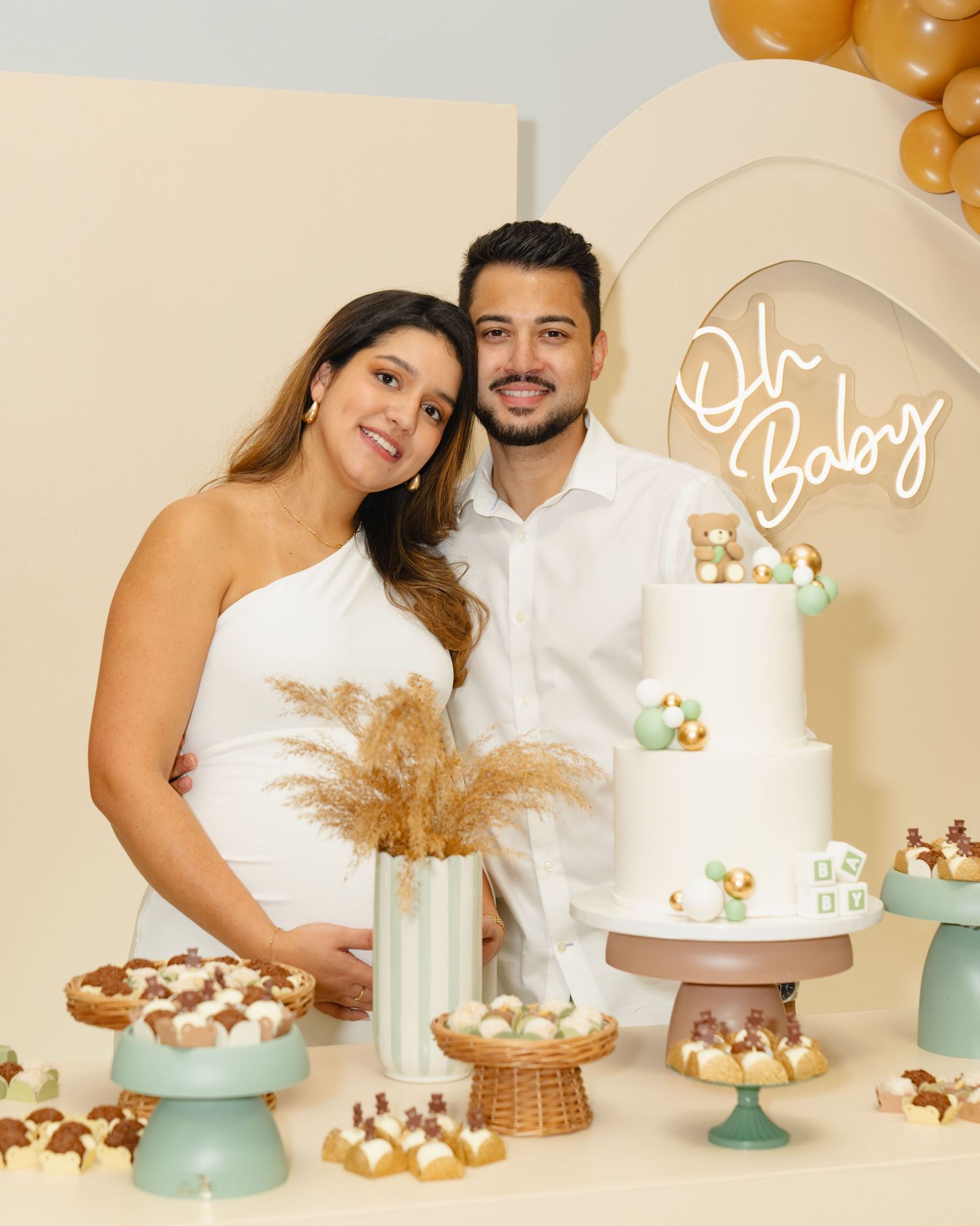 A couple poses by an elegant dessert display with a white tiered cake, pastries, and gold balloon decorations. Boston MA photographer.