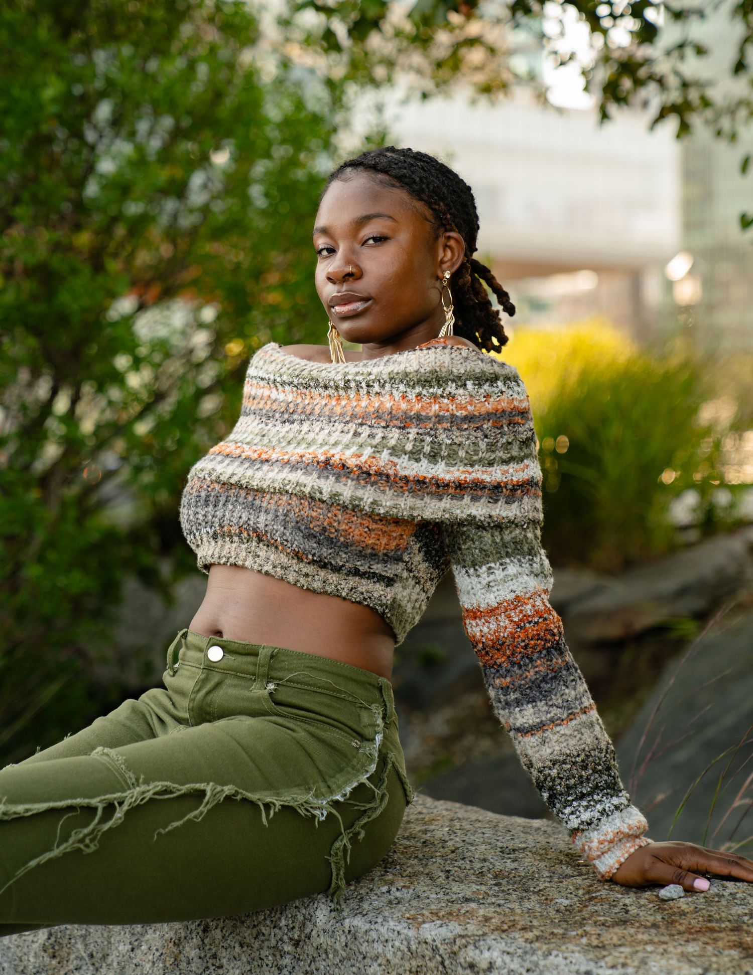 A person wearing olive green pants and striped knit sweater poses on a rock formation in a natural outdoor setting. Boston MA photographer.
