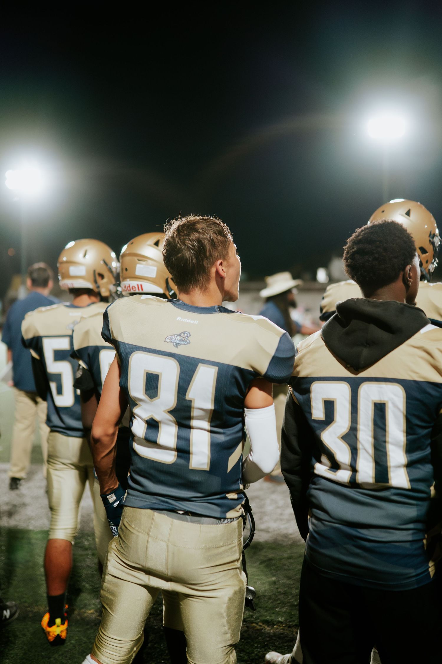 Football players in navy and white uniforms stand on sideline under bright stadium lights during nighttime game. Boston MA photographer.