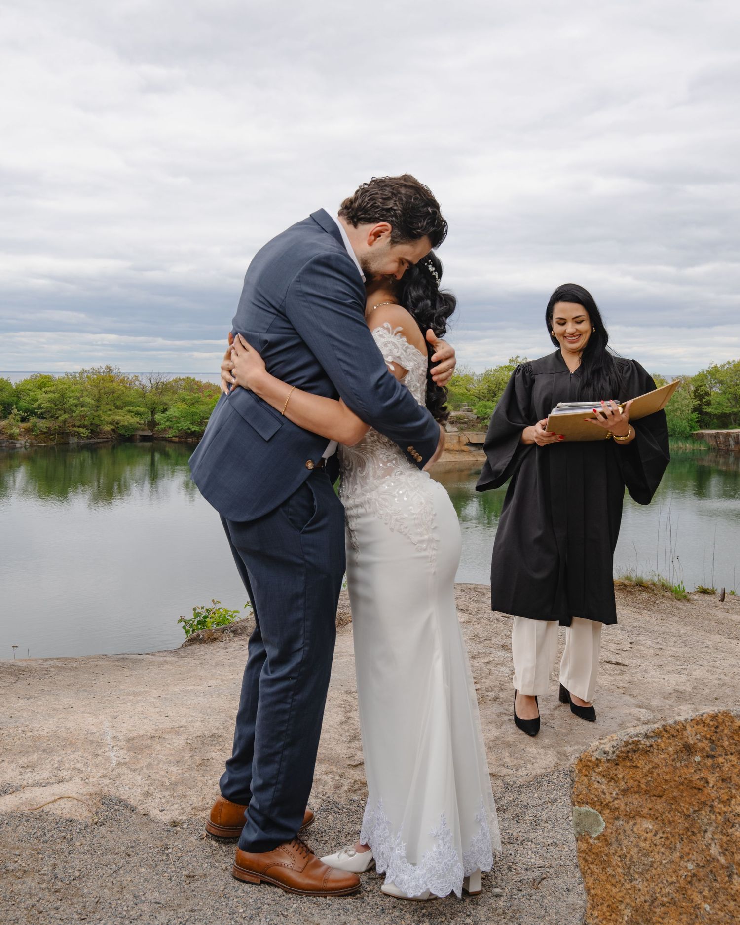 A couple embraces during their lakeside wedding ceremony while an officiant stands nearby. Boston MA photographer, Malden MA.
