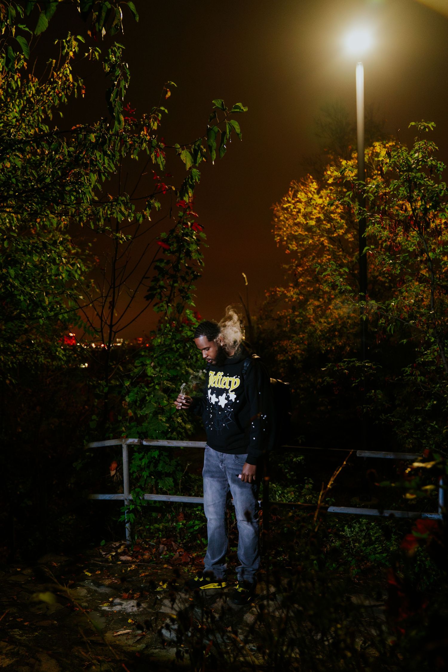 A person stands under a bright streetlight at night among dark trees and bushes in a park setting. Boston MA photographer, Malden MA.
