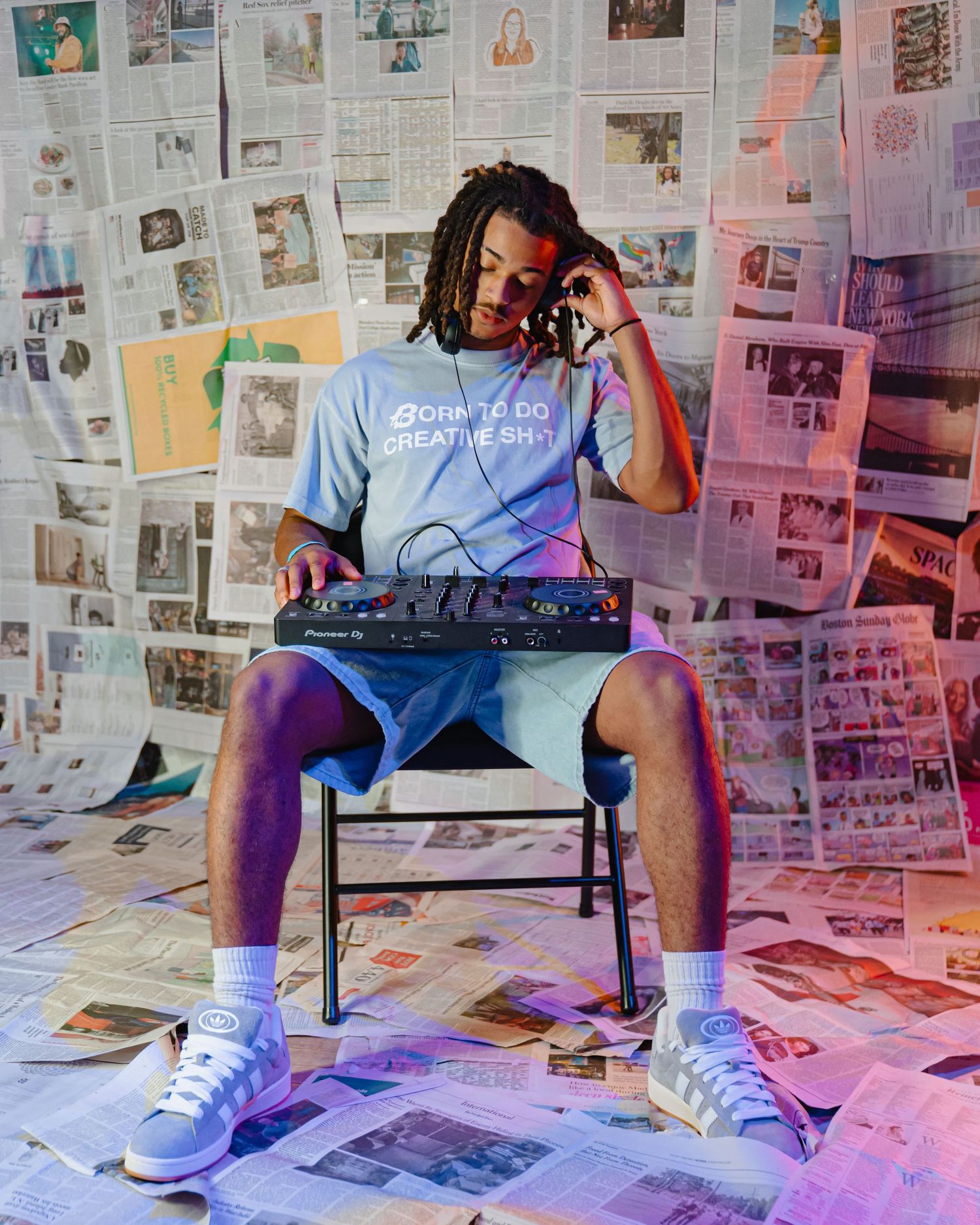 A person in blue shorts sits on a chair against a wall covered in newspaper clippings with colorful lighting effects. Boston MA photographer
