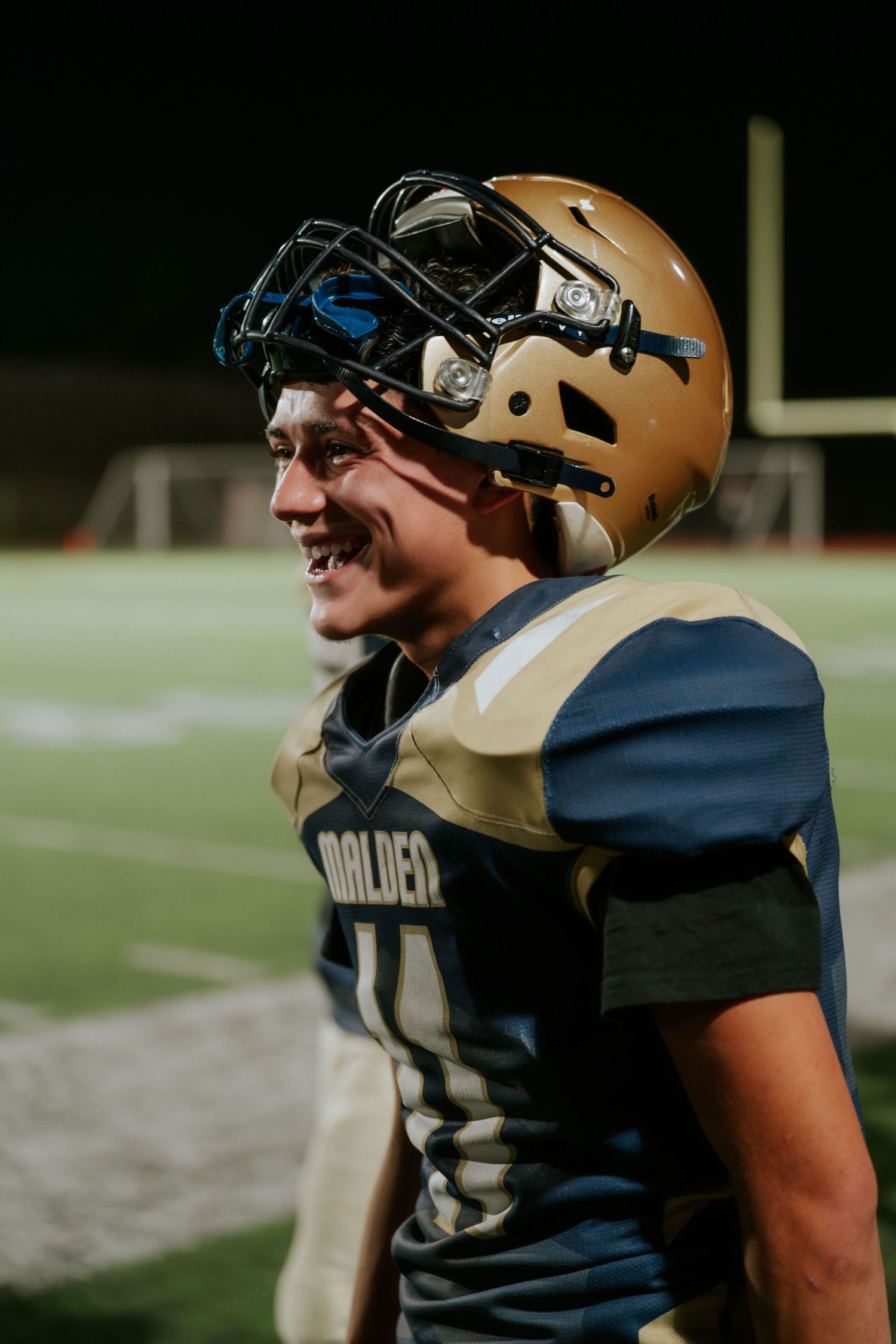 Football player wearing a gold helmet and navy uniform smiles while looking off to the side during a game. Boston MA photographer, Malden MA