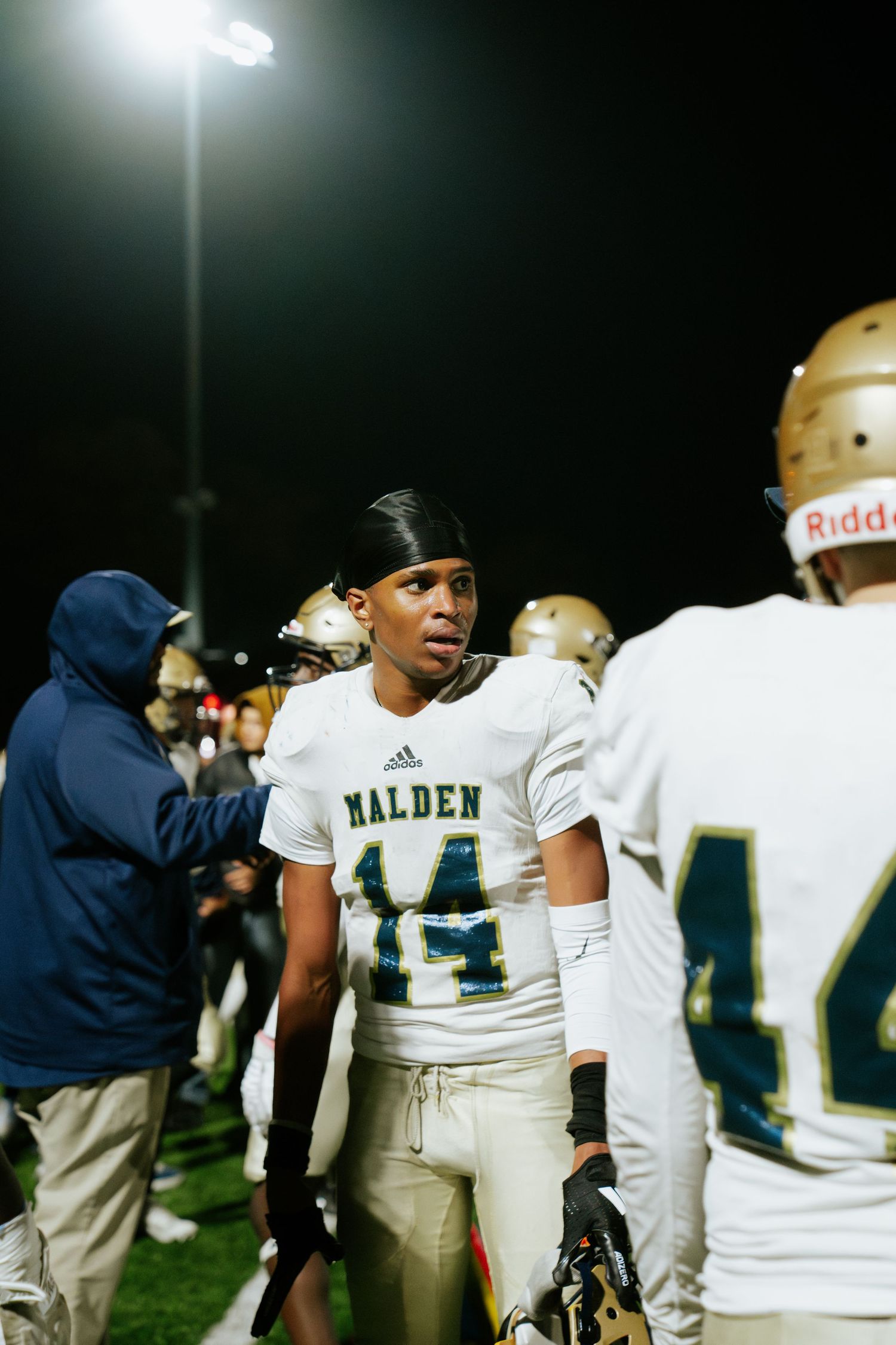 High school football players in white uniforms with green numbers stand on the sidelines during a night game. Boston MA photographer.