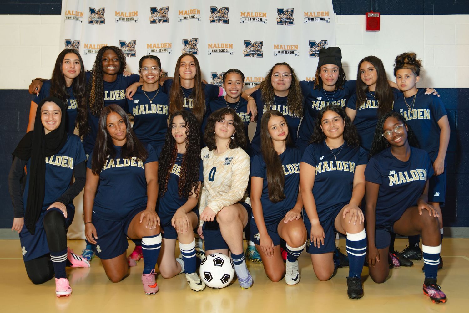A sports team poses together in matching navy blue uniforms with a soccer ball and basketball on the floor. Boston MA photographer.