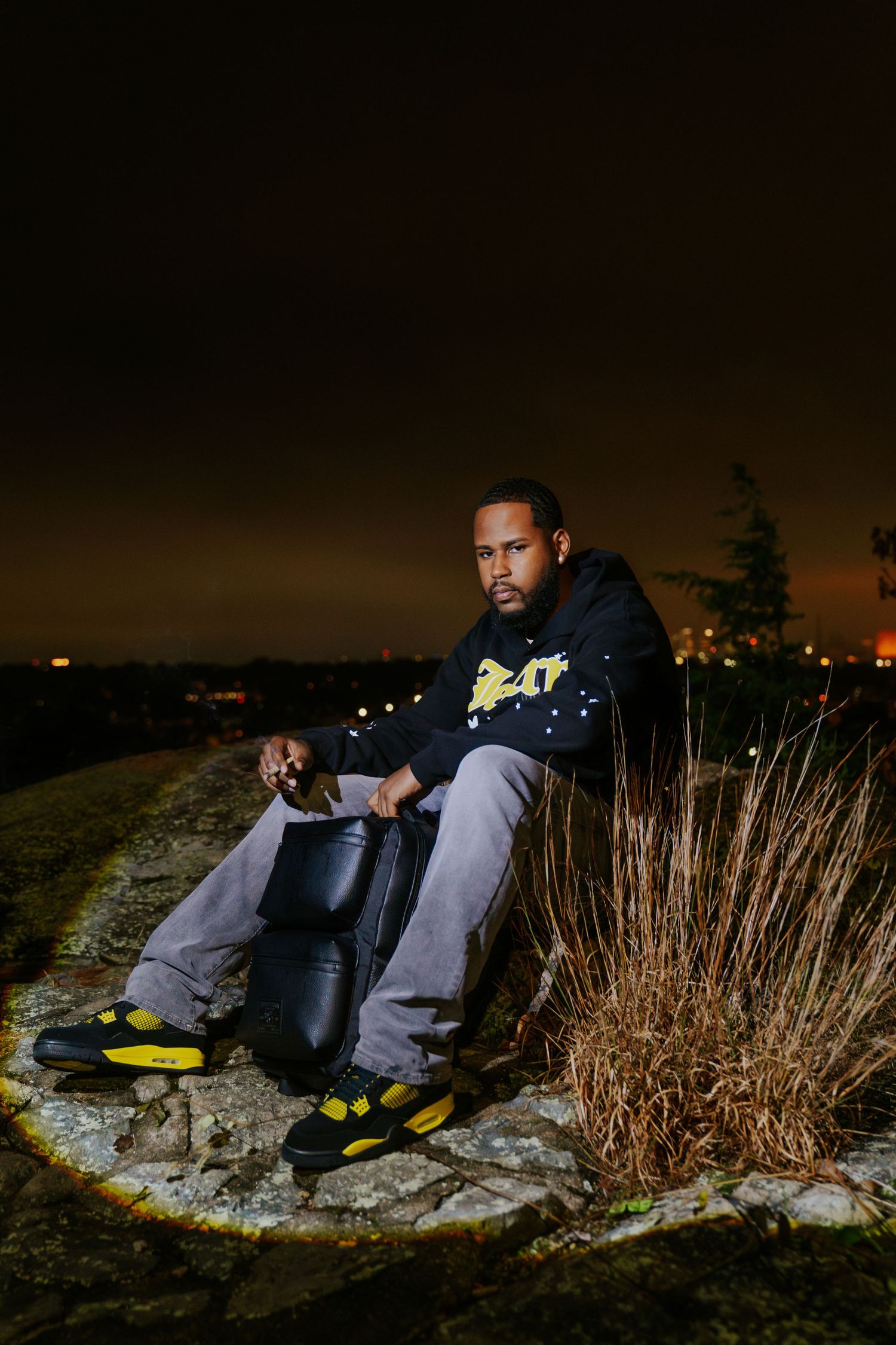 Person sitting outdoors at night on rocks wearing black jacket and grey pants with yellow accents in dark moody setting. Boston photographer