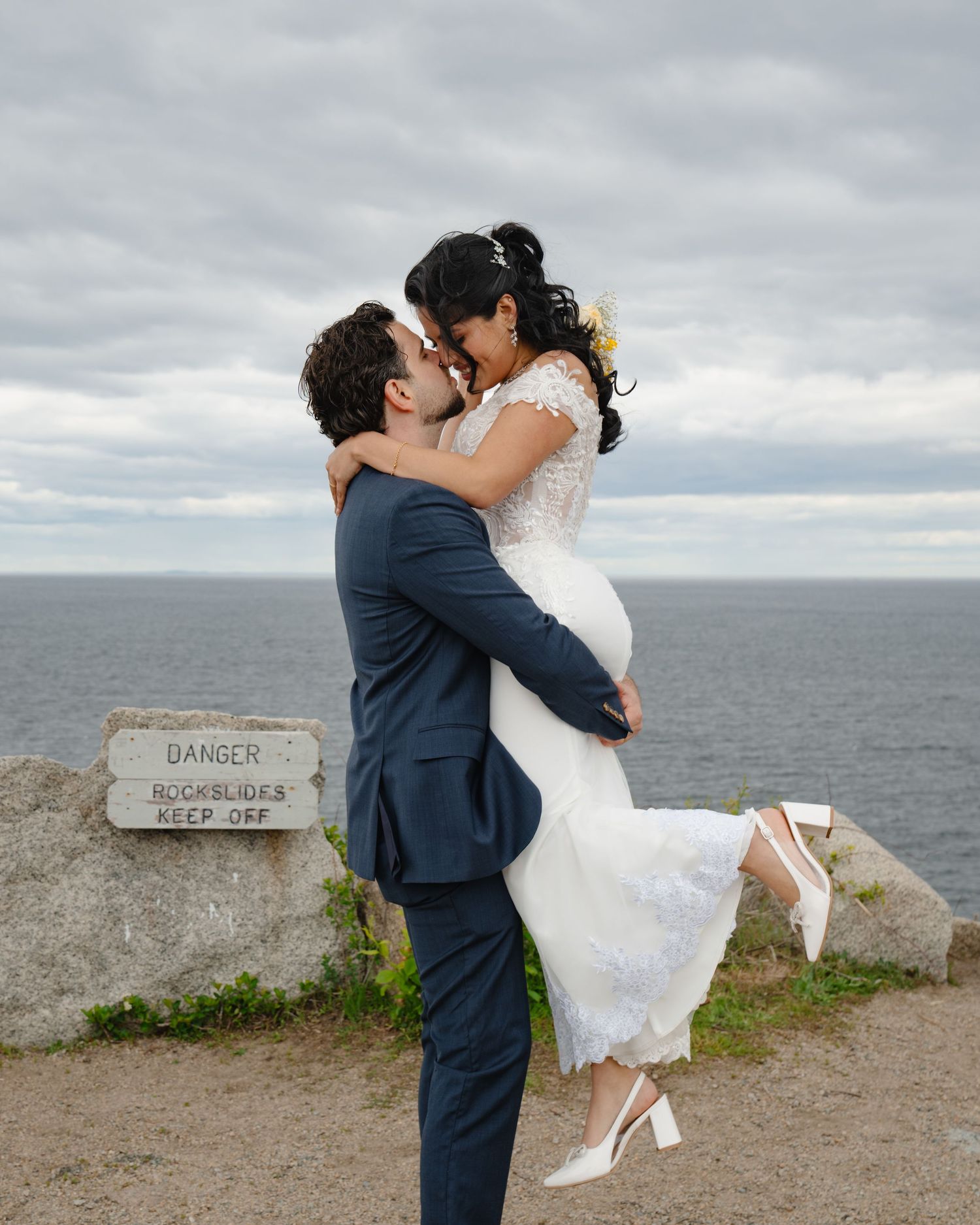 A couple in wedding attire embrace near a seaside cliff edge with dramatic cloudy skies in the background. Boston MA photographer.