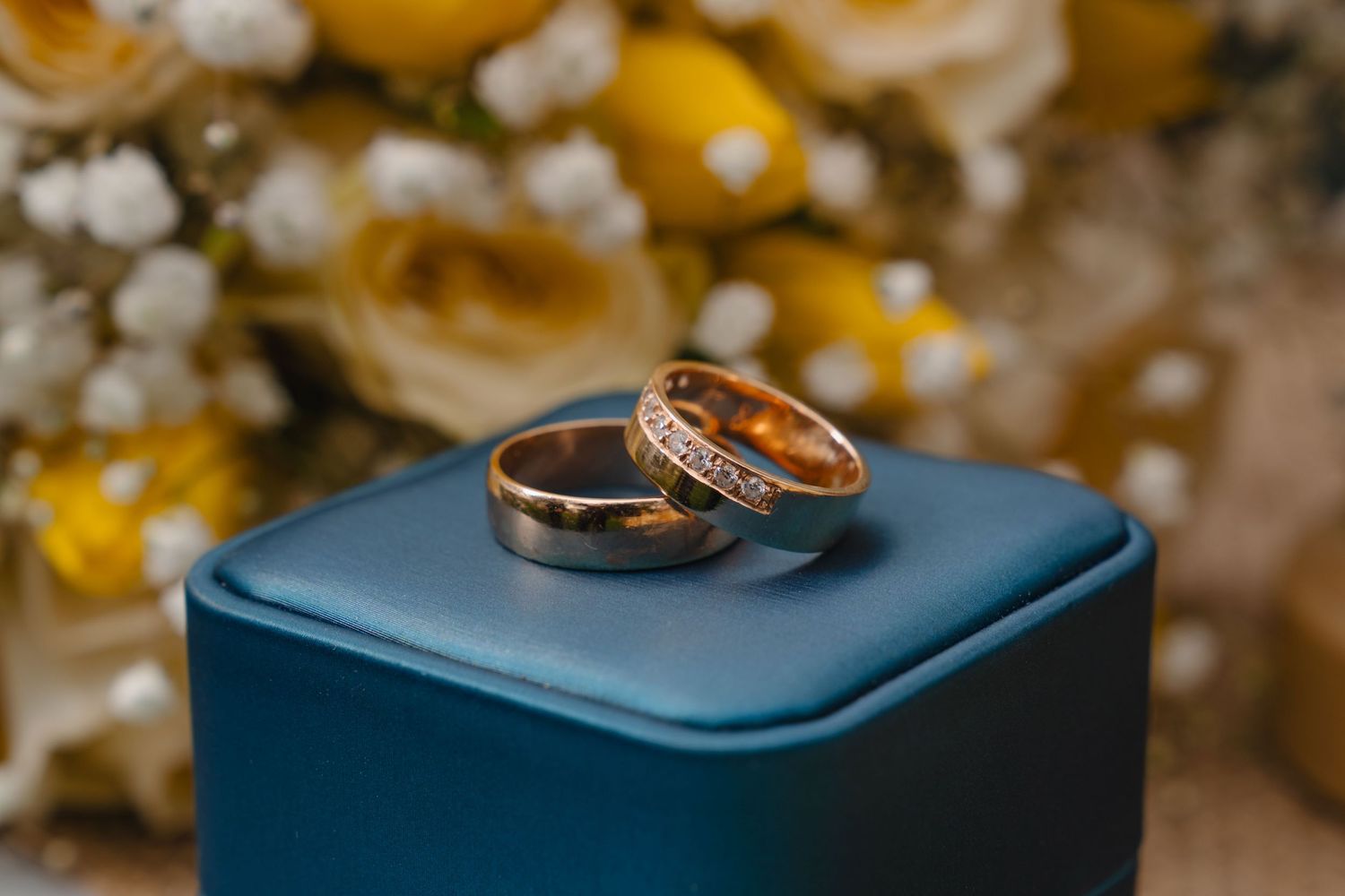 Wedding rings displayed on a navy blue ring box with blurred white floral decorations in the background. Boston MA photographer, Malden MA.