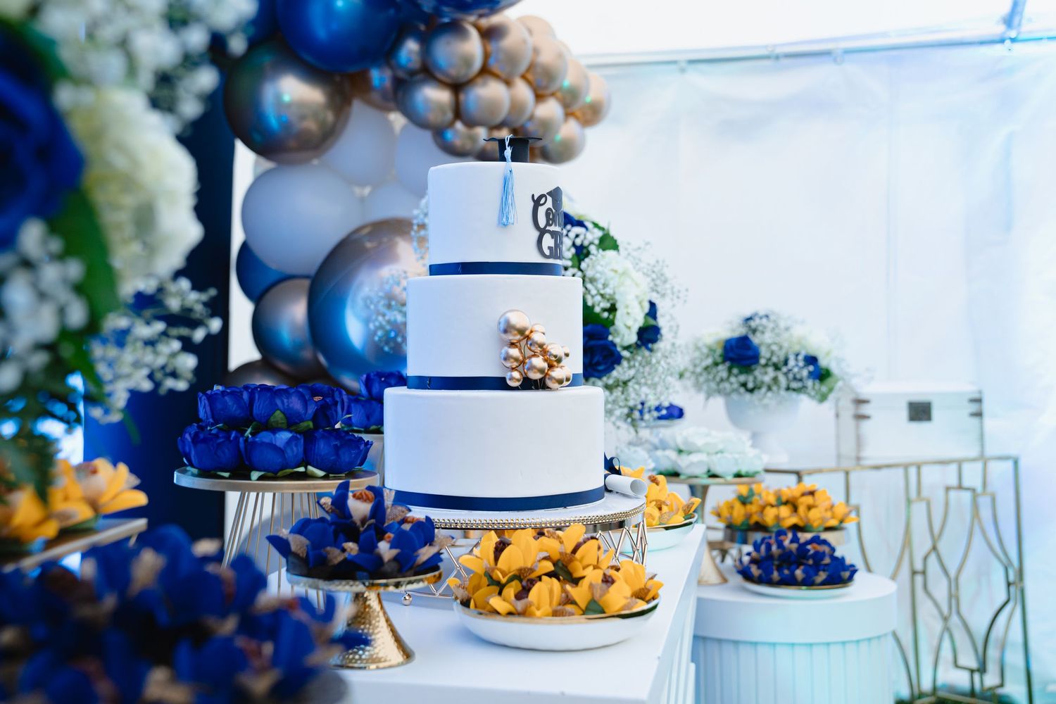 Elegant white wedding cake with blue and gold decorations displayed alongside dessert table at graduation party. Boston MA photographer.