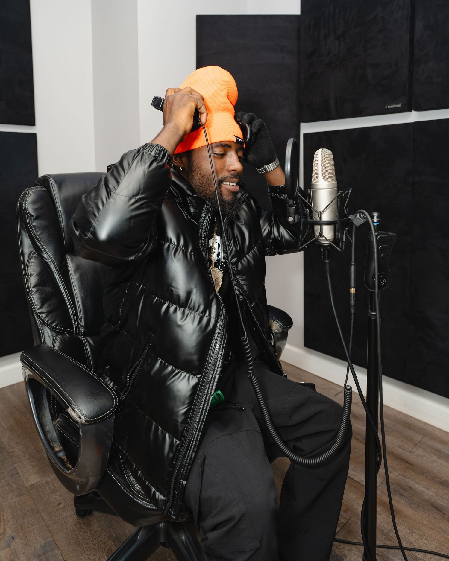 Person in a black puffer jacket and orange beanie recording vocals at a microphone in a studio setting. Boston MA photographer, Malden MA.