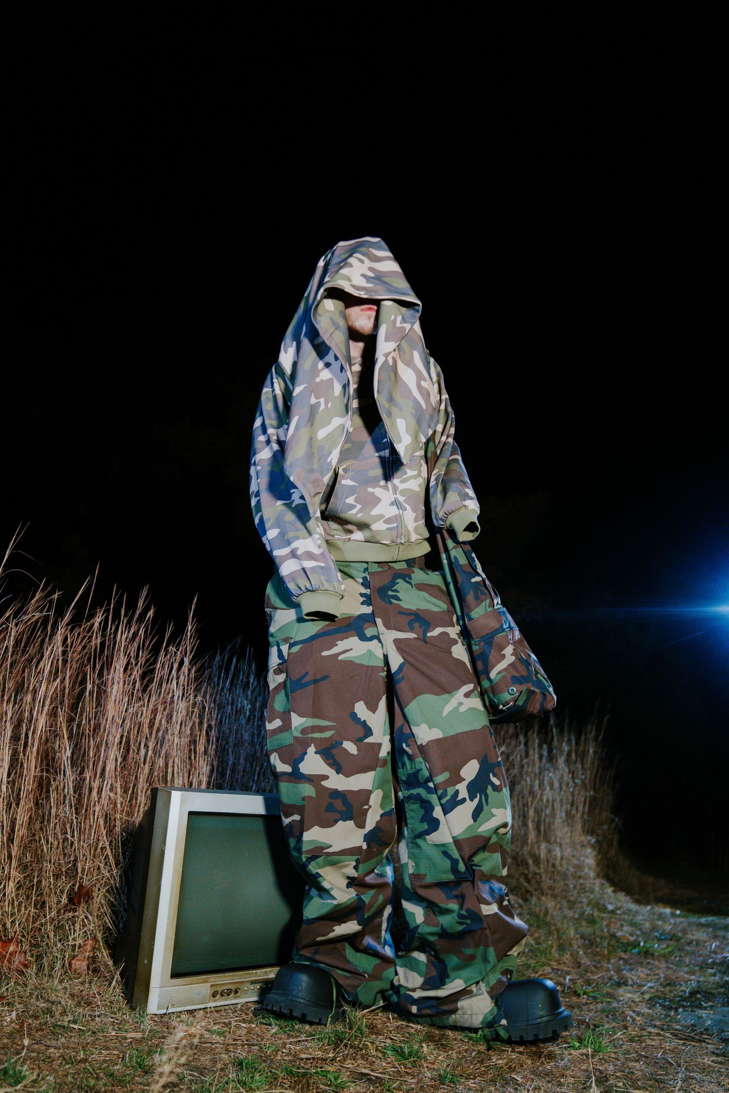 Military camouflage backpack rests against tall grass and a green box in a dark outdoor setting. Boston MA photographer, Malden MA.