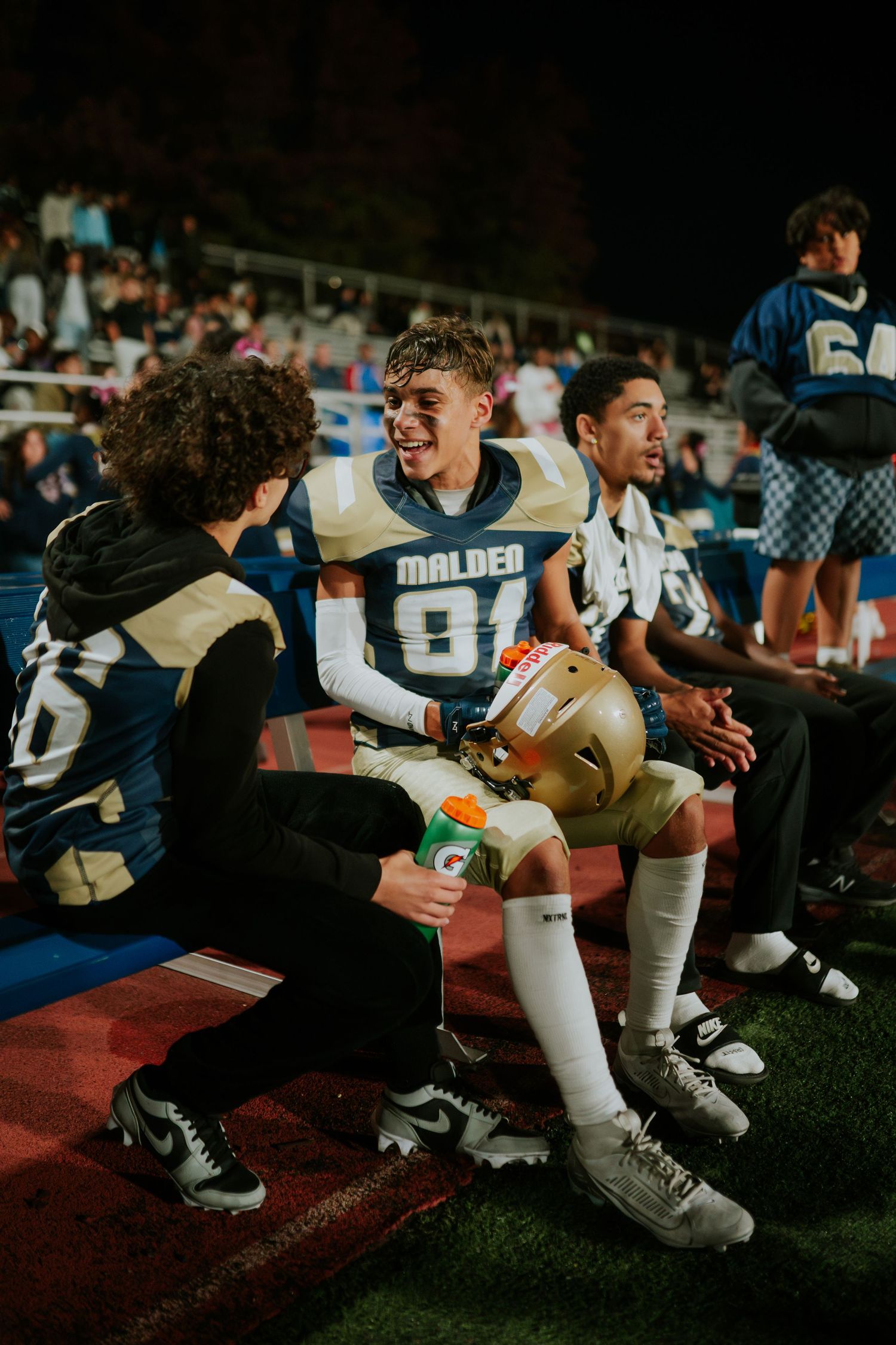 Athletes sitting on sidelines during a football game share a light-hearted moment together on the field. Boston MA photographer, Malden MA.
