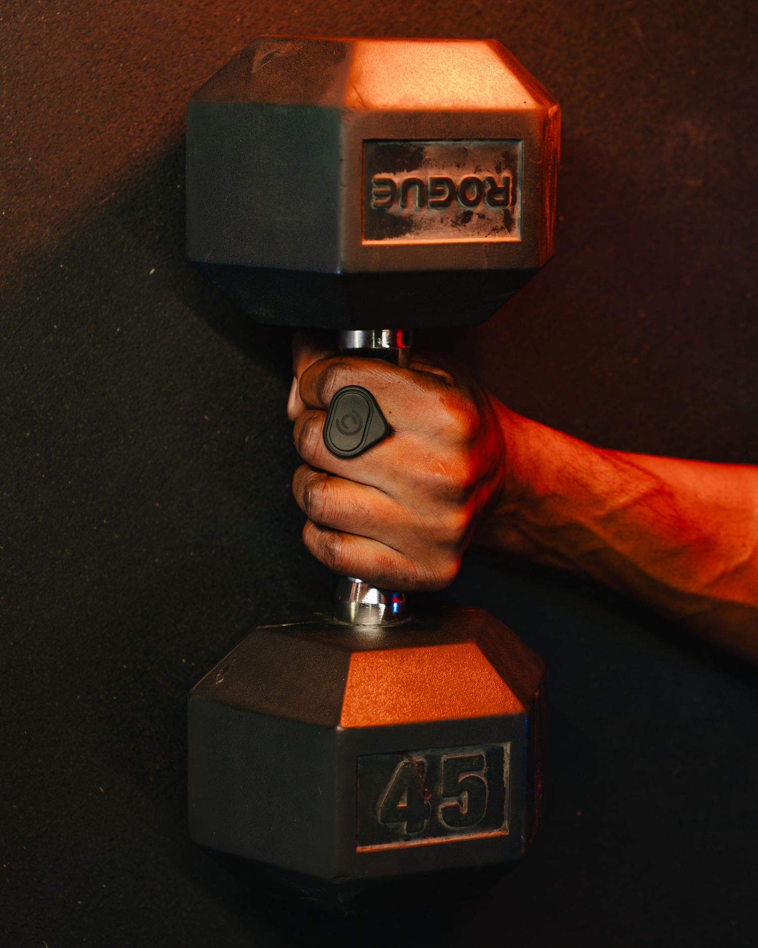 A hand grips a black 45-pound dumbbell against a dark background with dramatic orange lighting. Boston MA photographer, Malden MA.
