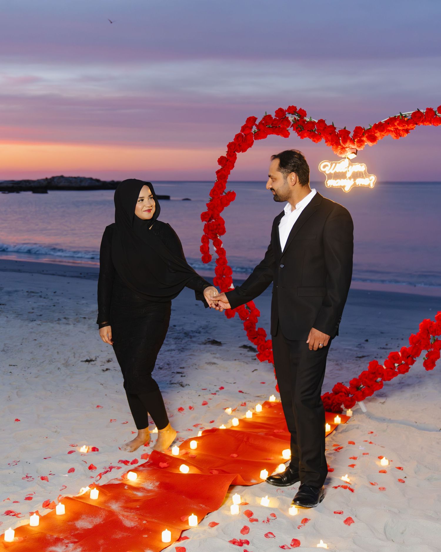 A romantic beach proposal scene at sunset with red heart-shaped rose petals and candlelit pathway on the sand. Boston MA photographer.