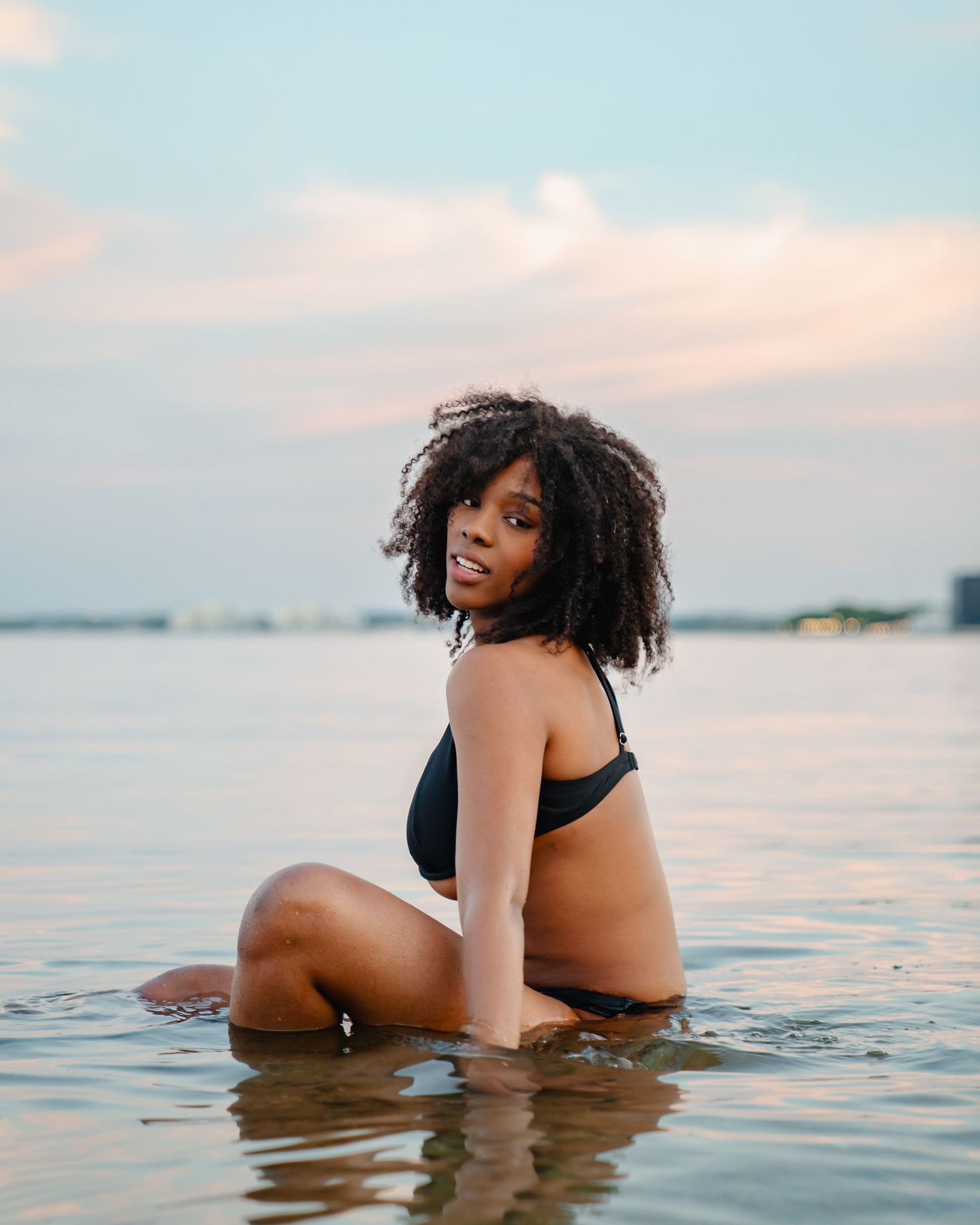 A person in a black swimsuit poses in shallow water at sunset with a natural curly hairstyle. Boston MA photographer, Malden MA.