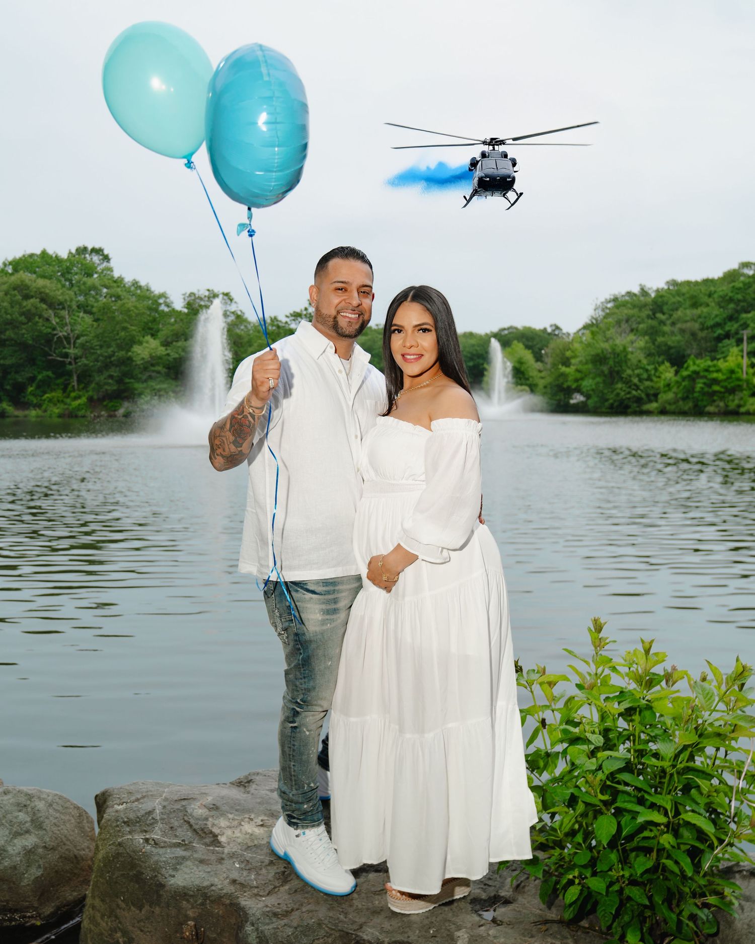 A couple poses by a lake with blue balloons. Helicopter flies overhead and water fountains spray in the background. Boston MA photographer.
