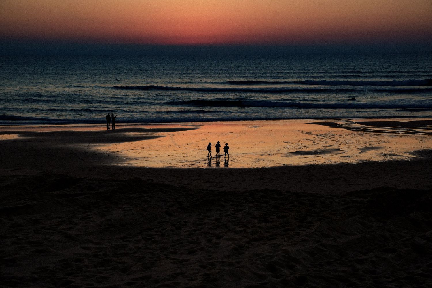Silhouetten von Kindern, die bei Sonnenuntergang am Strand in Frankreich spielen, während der Sonnenuntergang sich im nassen Sand spiegelt.