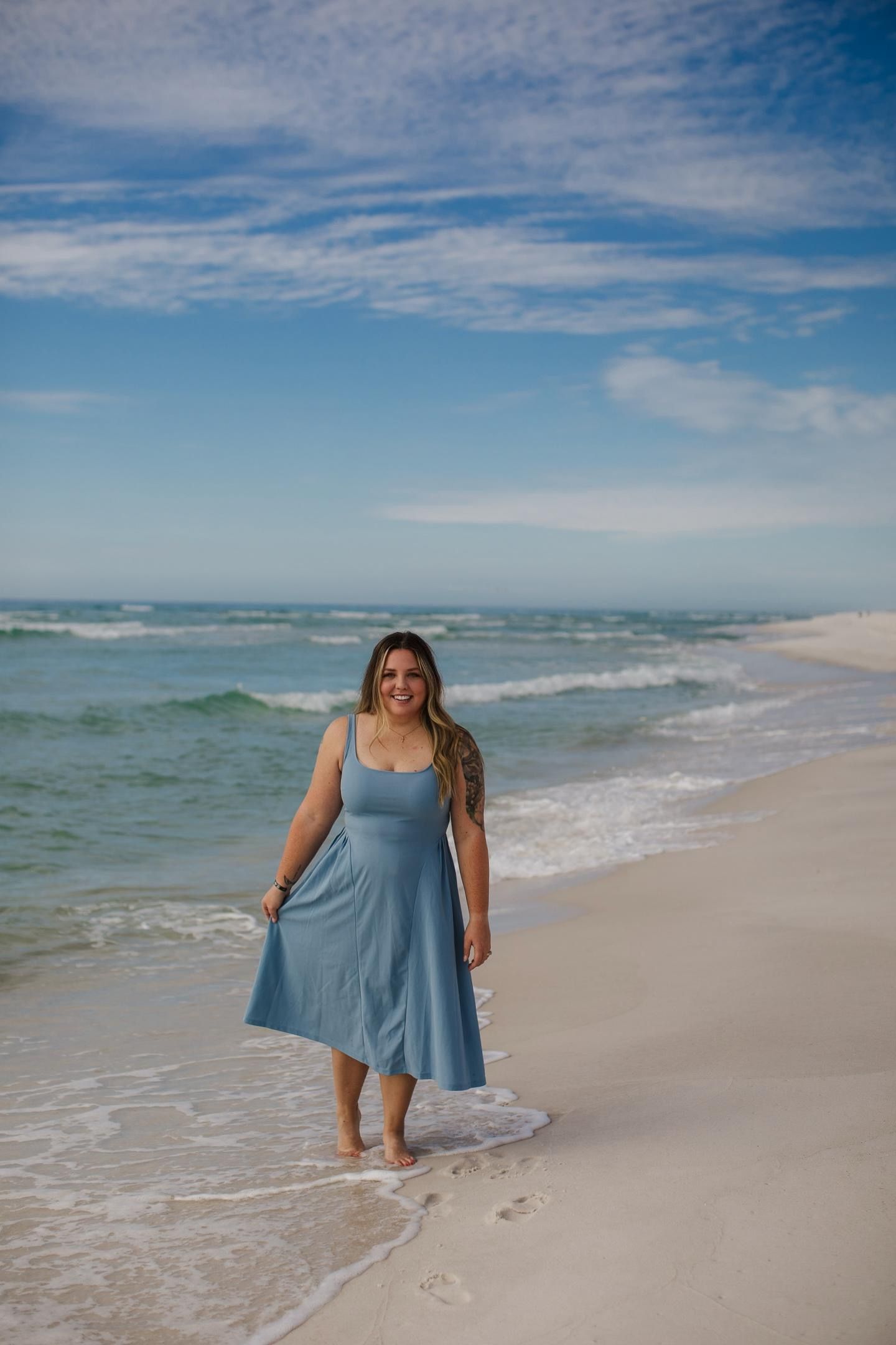 Woman in flowing blue dress twirls and dances along a pristine white sand beach with turquoise ocean waves.