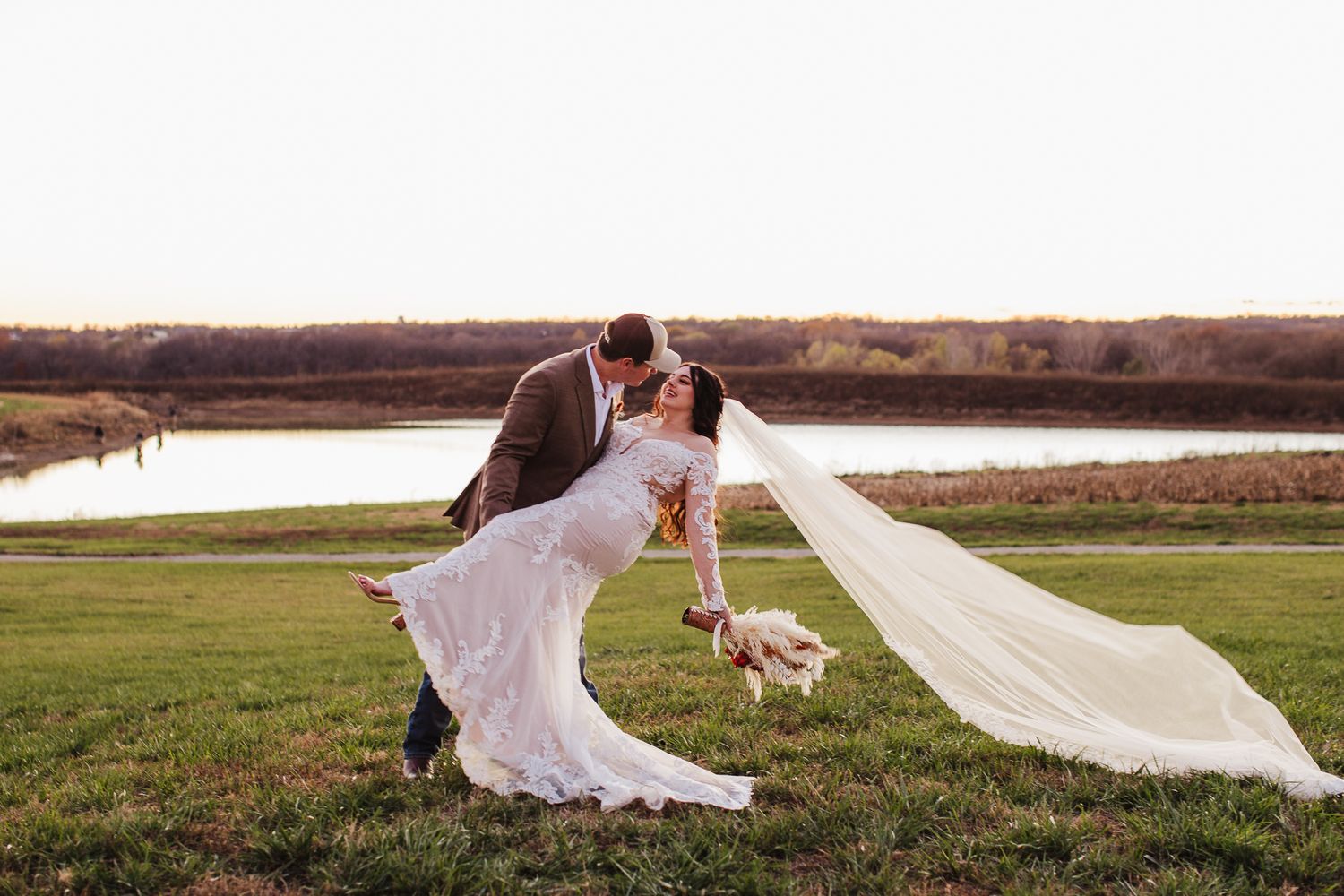 A romantic wedding moment captured on a grassy hill overlooking a serene lake with a flowing veil catching the breeze.