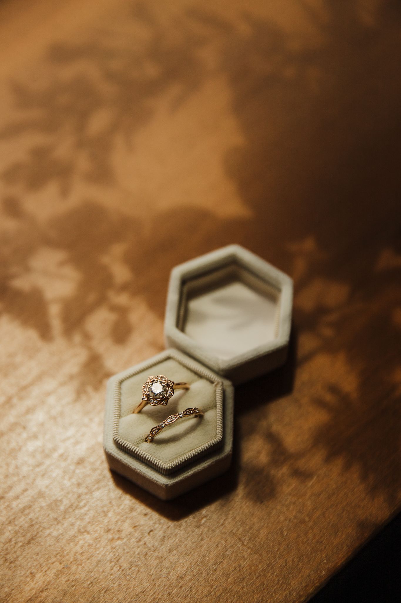 Diamond engagement ring displayed in a gray hexagonal box on wooden surface in warm lighting.