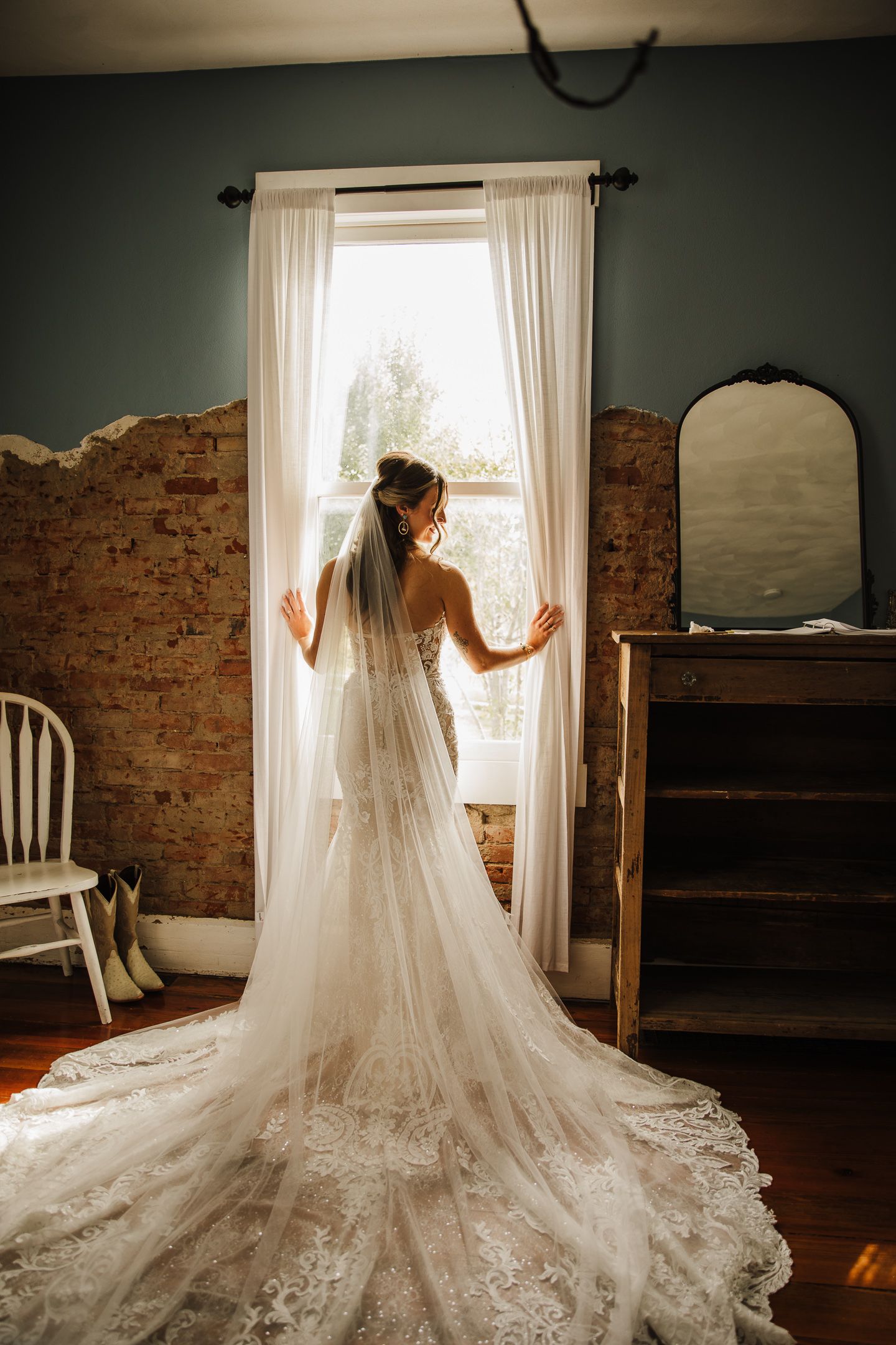 Silhouette of bride in lace gown standing at window with exposed brick wall and vintage decor.