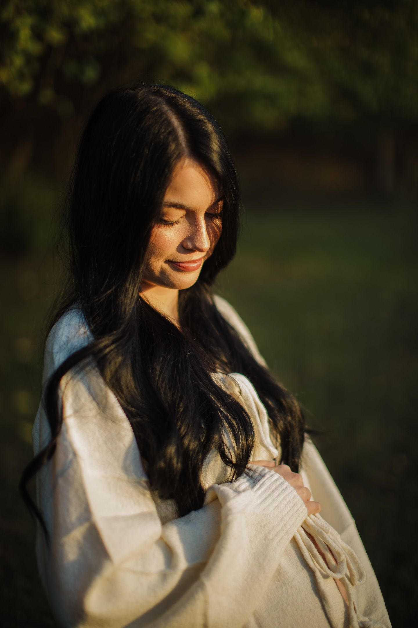 Woman in white sweater poses thoughtfully during golden hour maternity photoshoot.