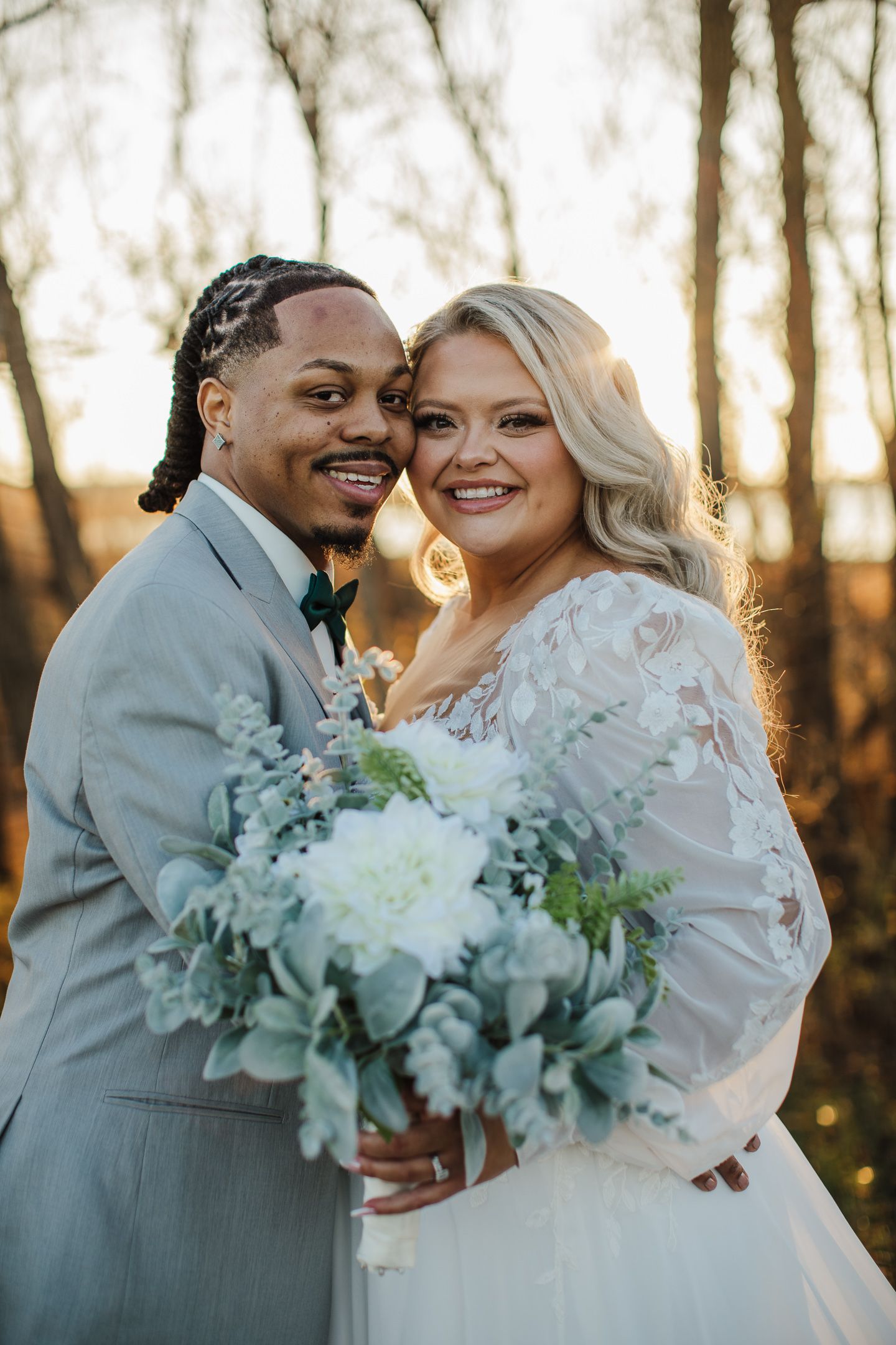 A newlywed couple poses with a white floral bouquet at sunset.