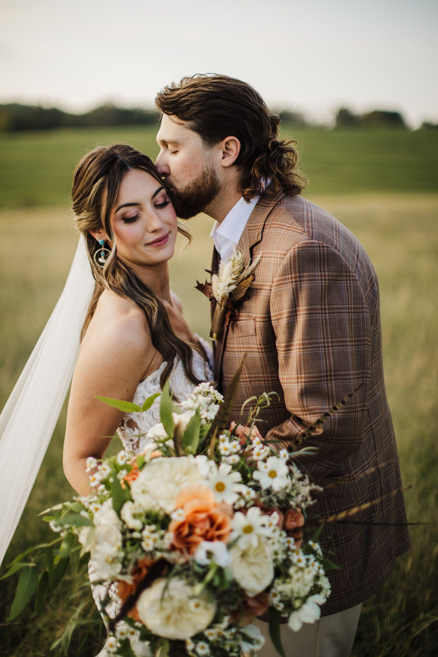 Romantic wedding couple embraces in golden field at sunset with rustic bouquet of wildflowers and white blooms.