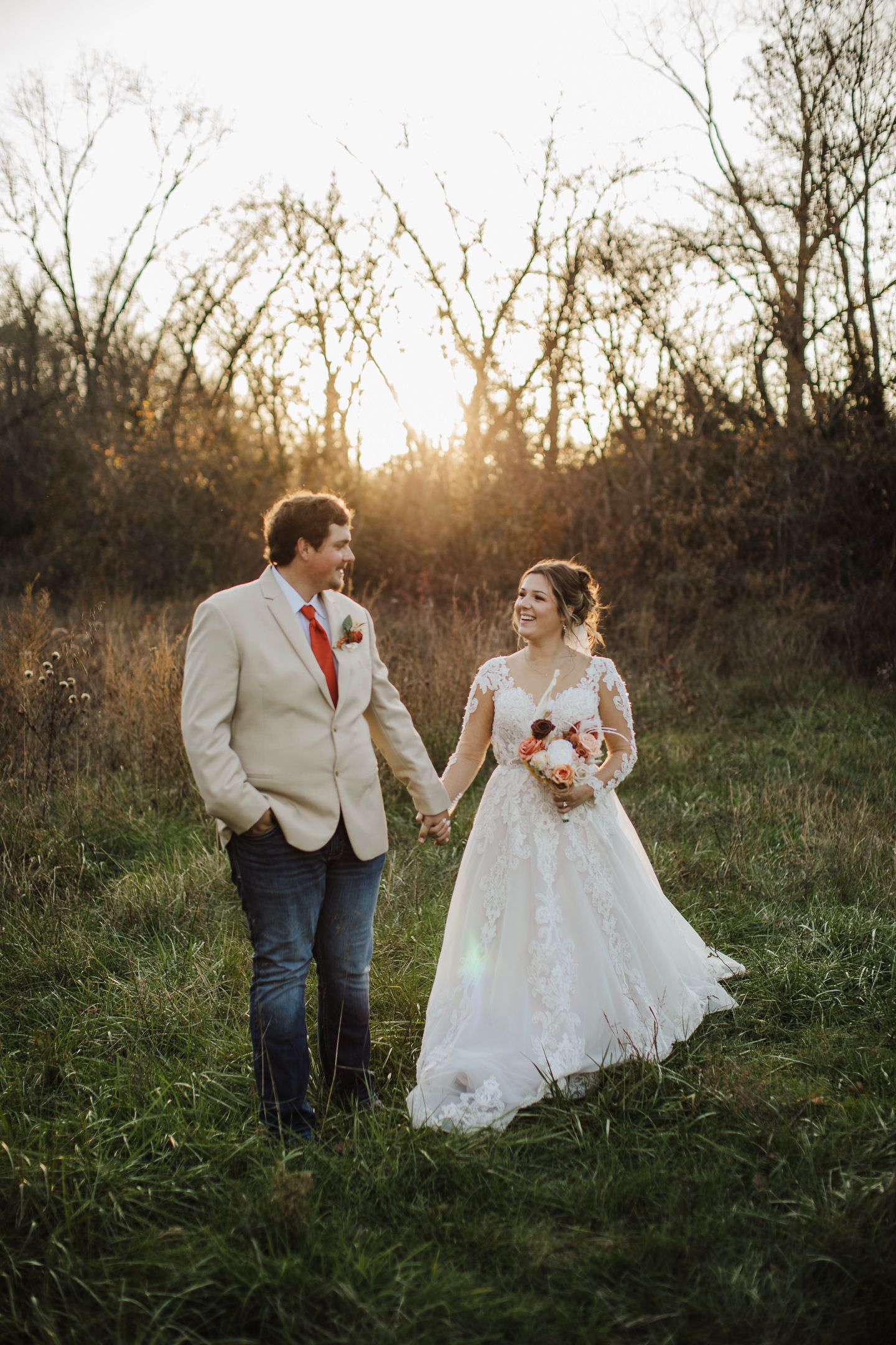 A couple stands together in a field at sunset with warm golden light filtering through trees in the background.