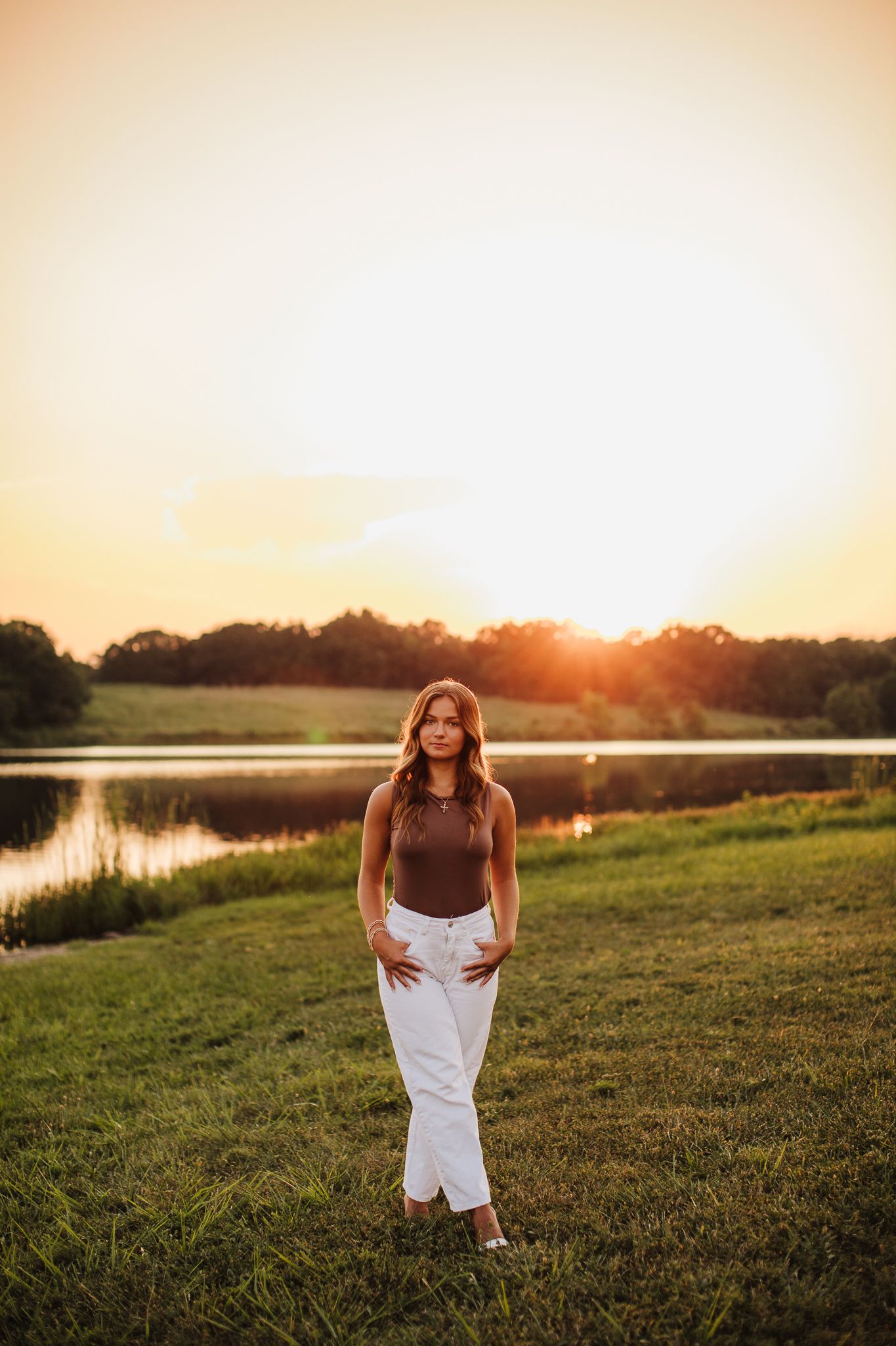 Person in white dress walks through grassy field at sunset near a lake.