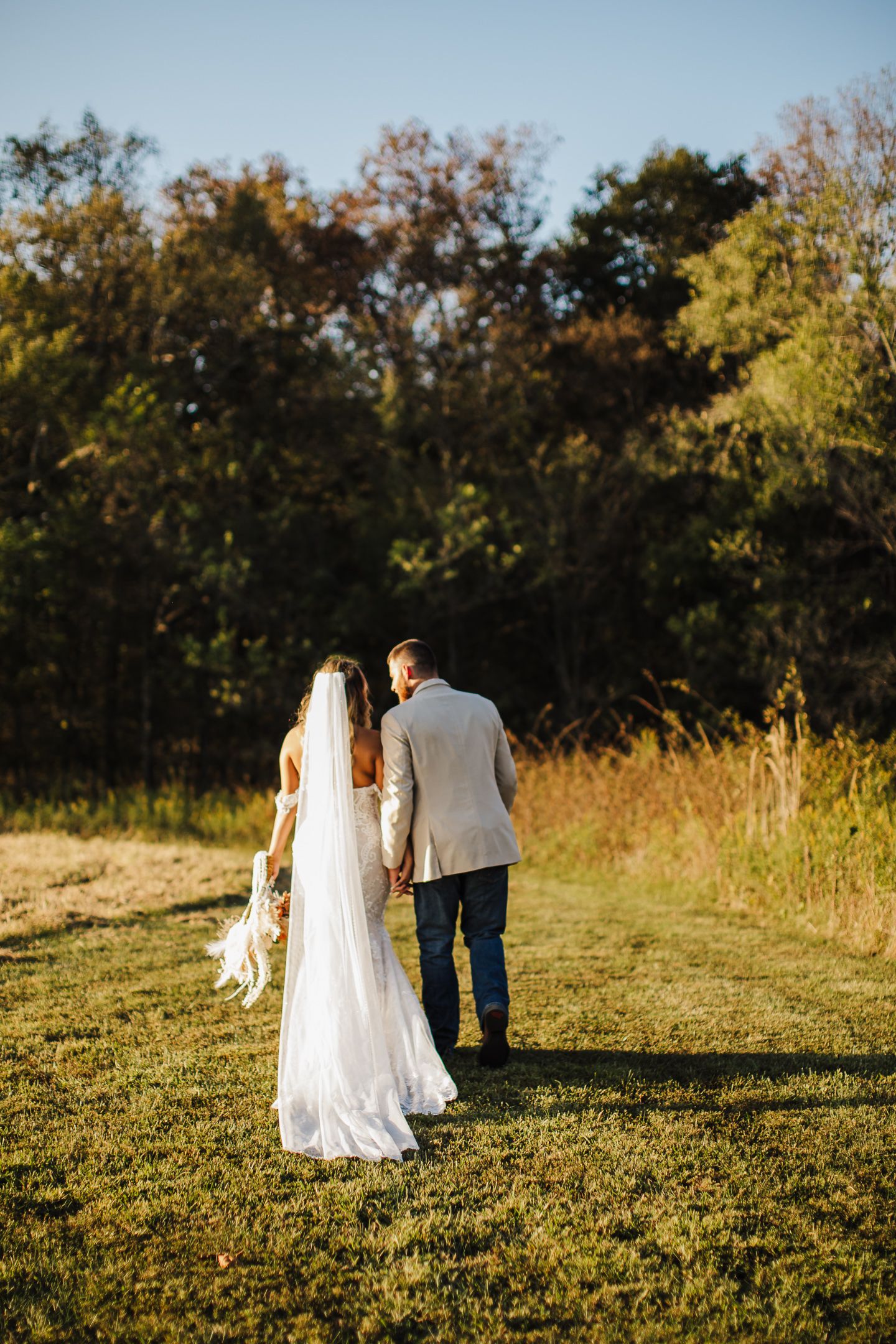 A bride and groom walk together through a sunlit meadow during golden hour as trees line the horizon.