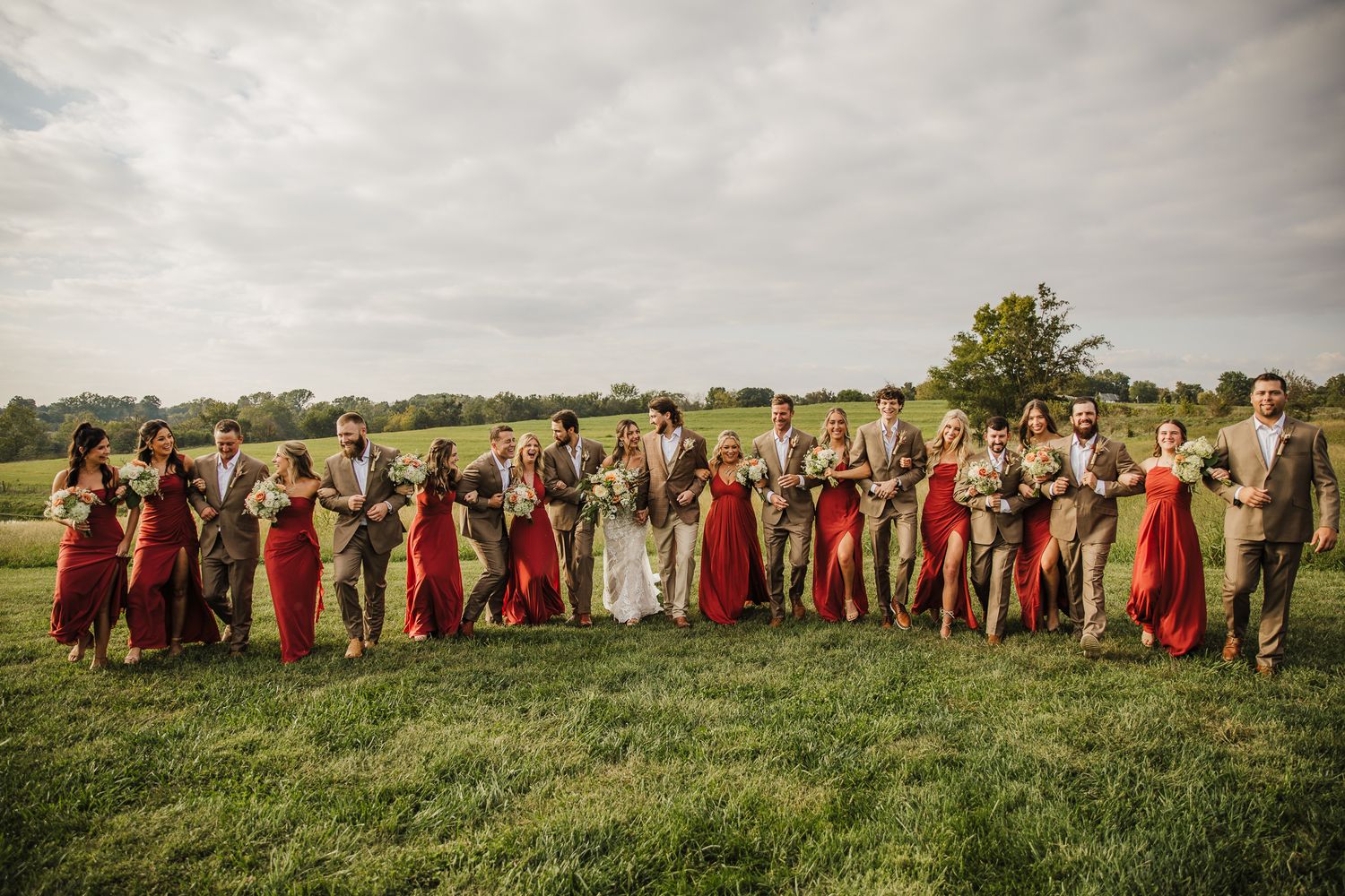 Large wedding party in red and neutral colors poses together in a scenic outdoor field.
