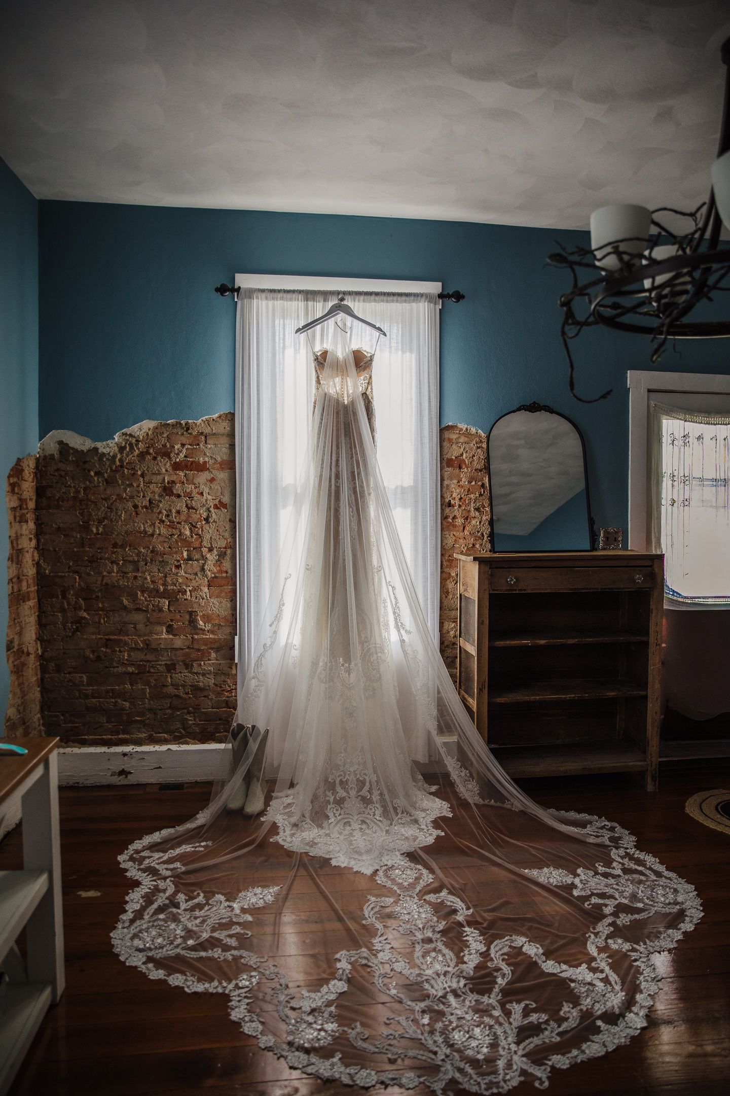 Elegant lace wedding dress with long train displayed against exposed brick walls and teal colored room.