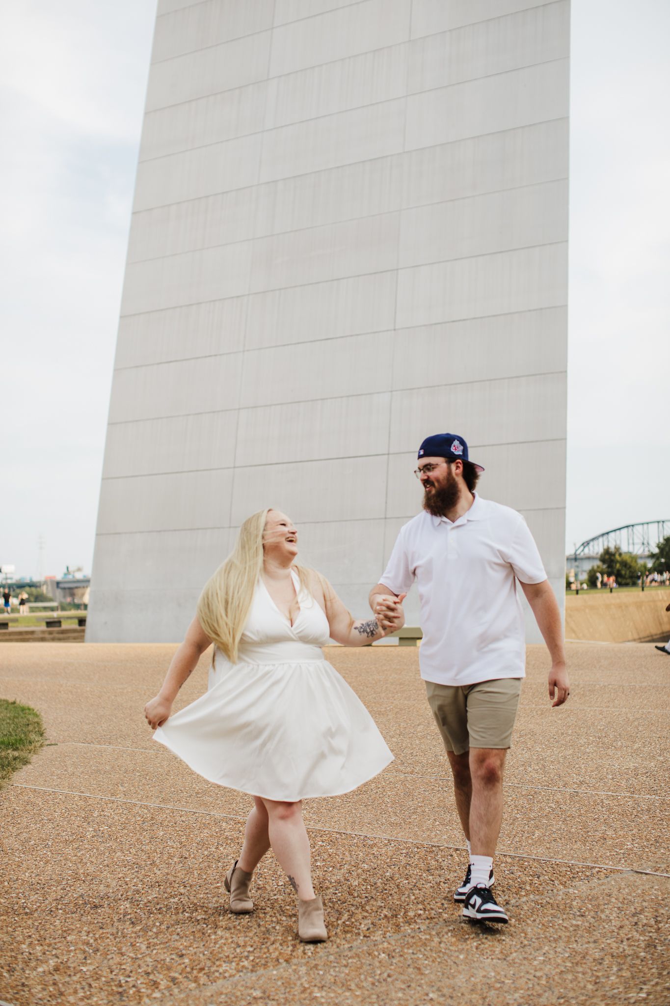 Playful couple in white outfits dancing and twirling against a light colored wall outdoors.
