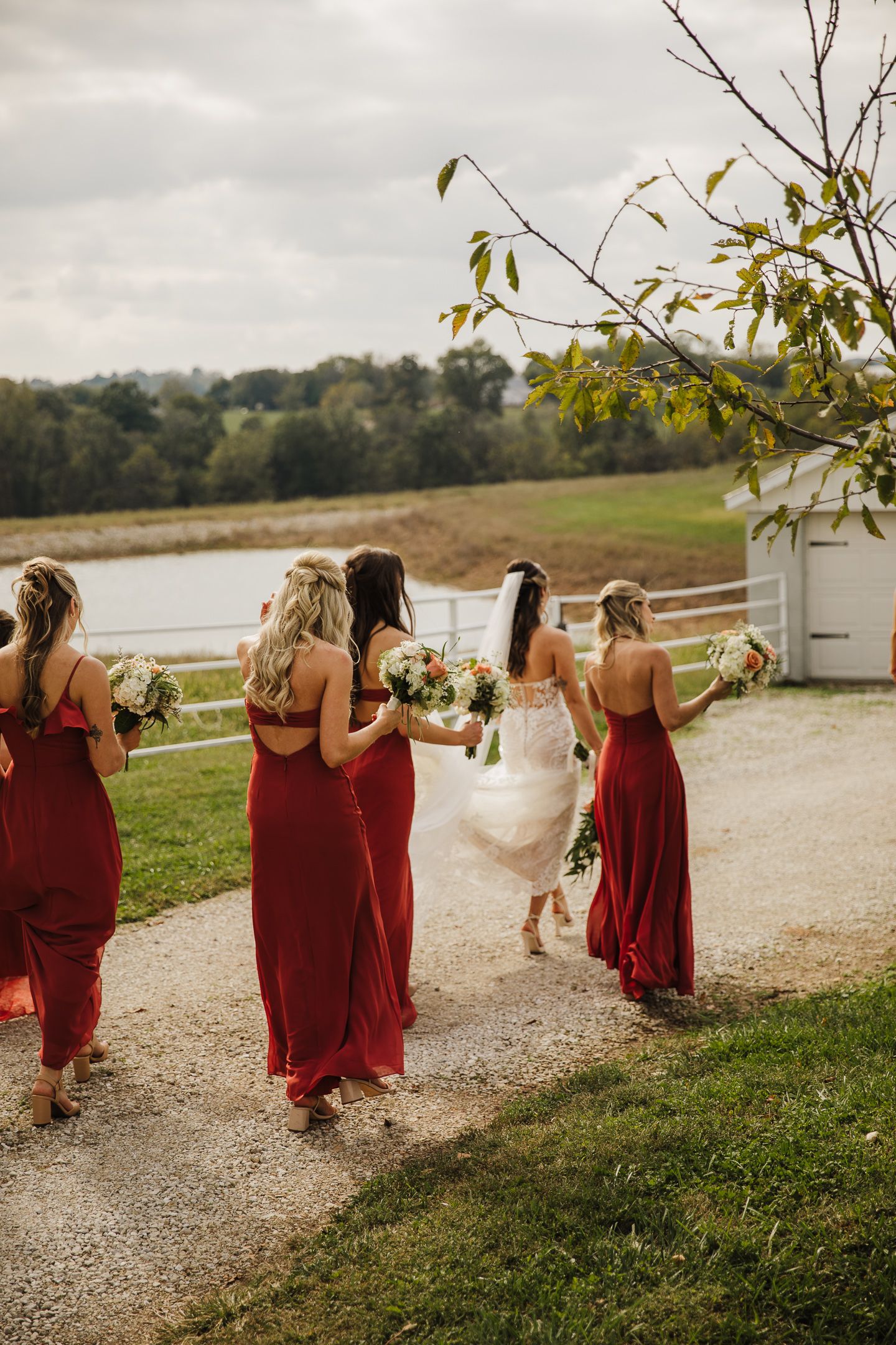 Bridesmaids in burgundy dresses walk along a lakeside path during an autumn wedding.