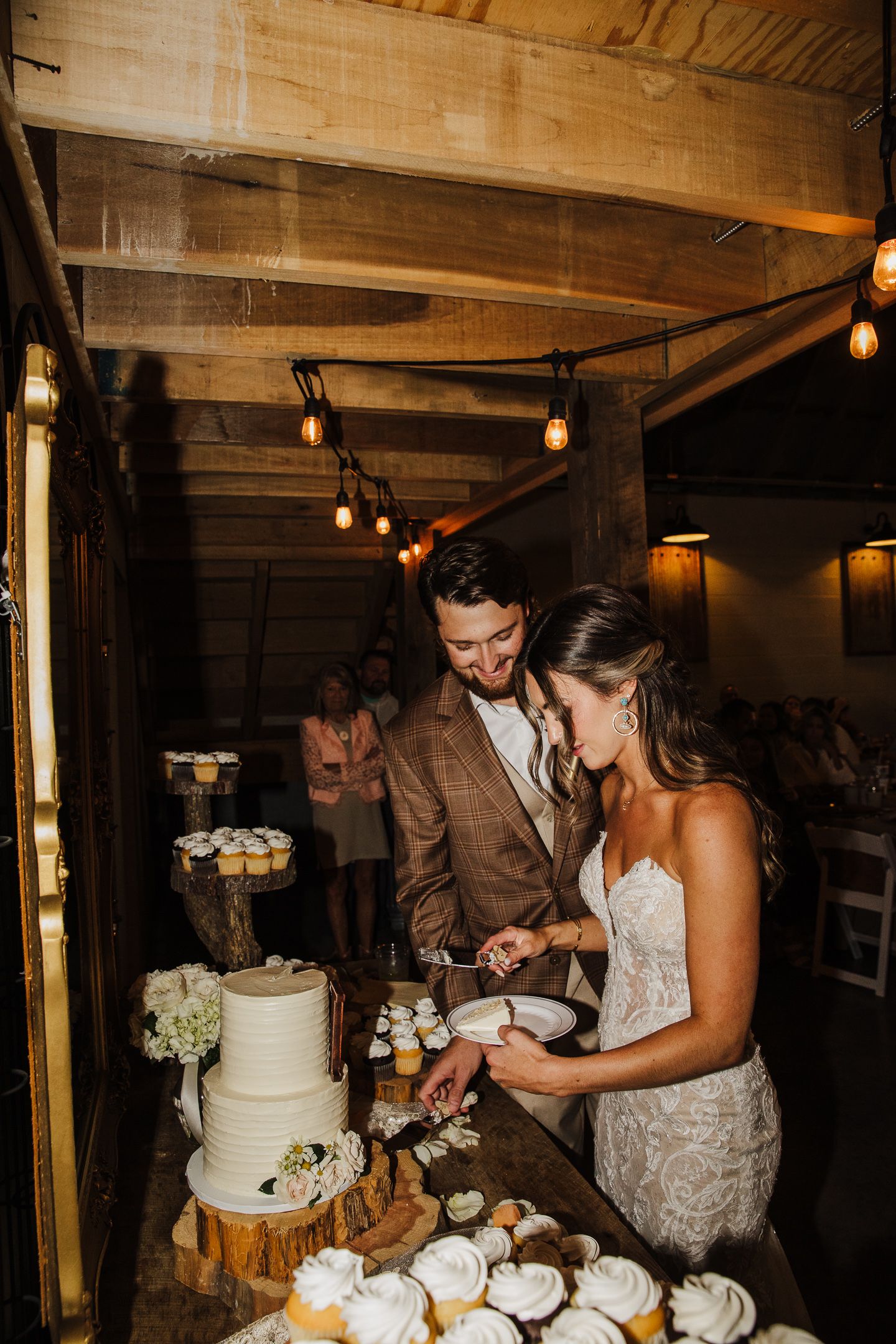 Wedding couple cutting their elegant white tiered cake in rustic venue with string lights.